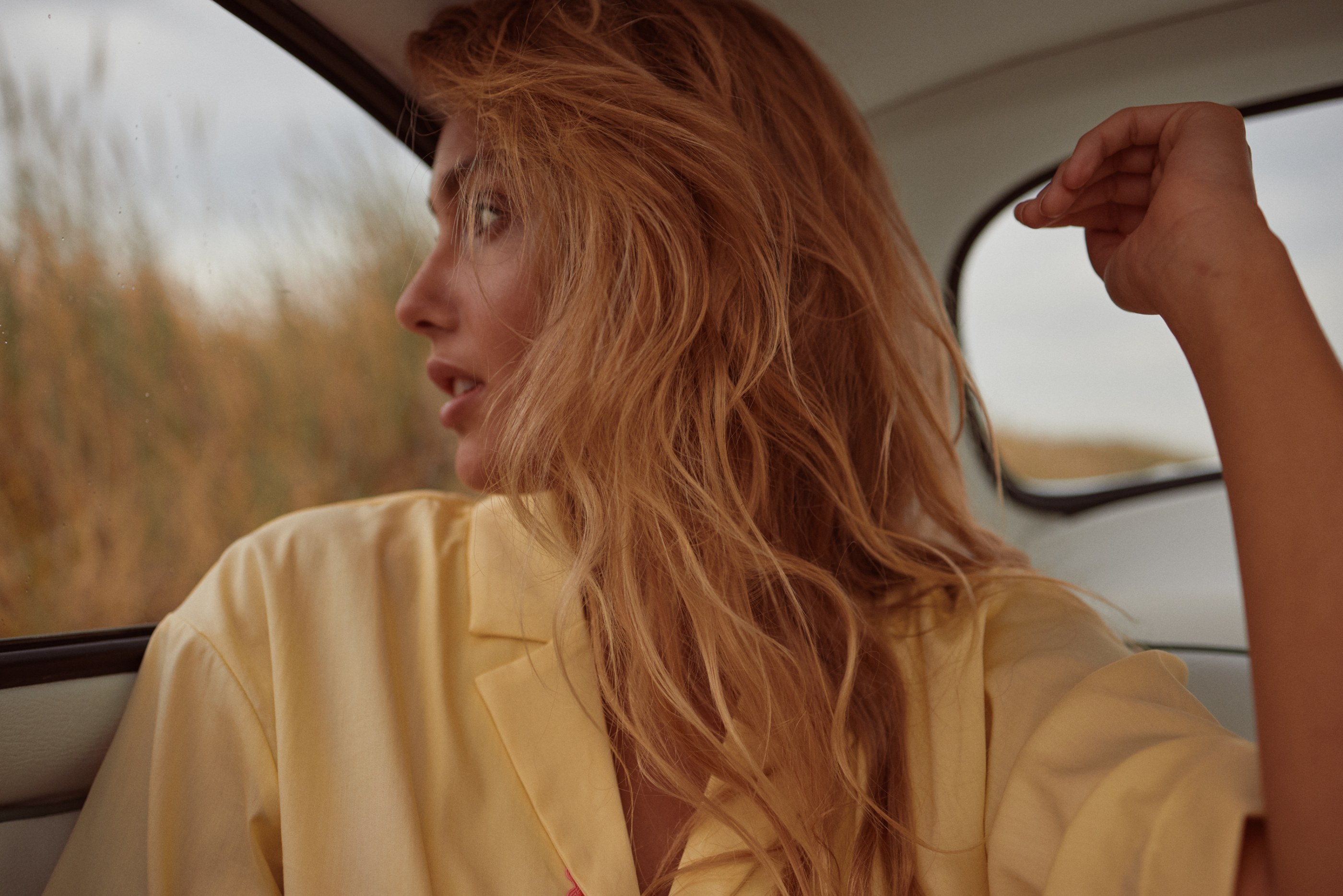 Woman with red hair in a yellow blazer looking out a car window at a blurry field.