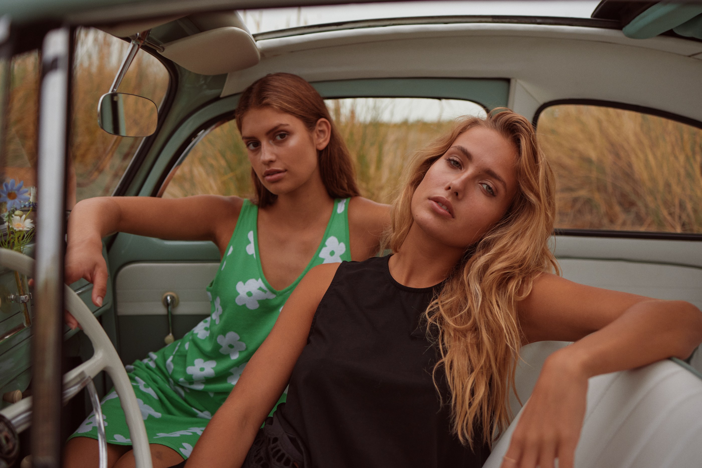 Brown-haired woman in green floral dress, blonde woman in black top, sitting in vintage car.