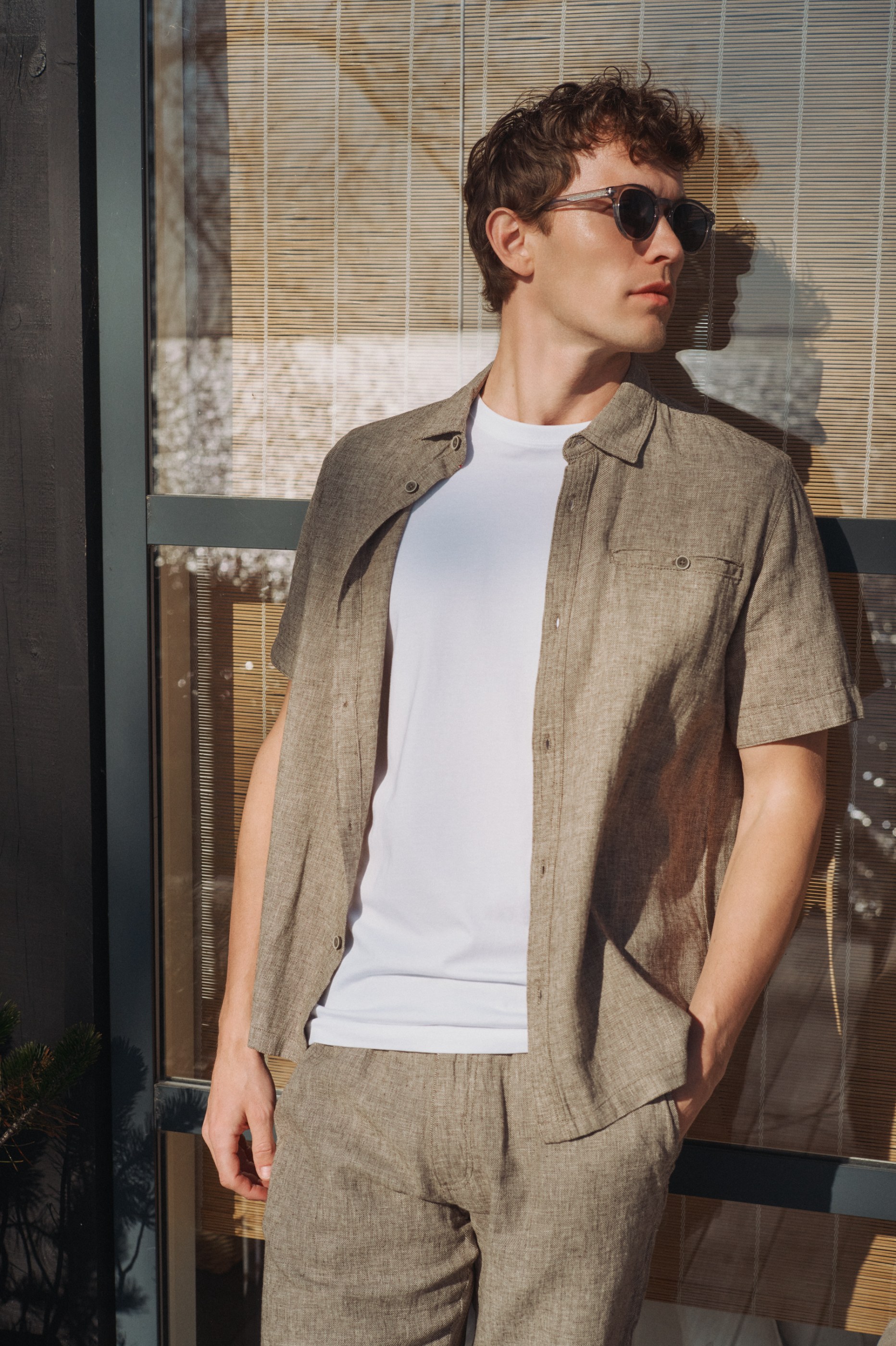 Man in sunglasses and brown linen outfit stands by a sunlit bamboo blind.