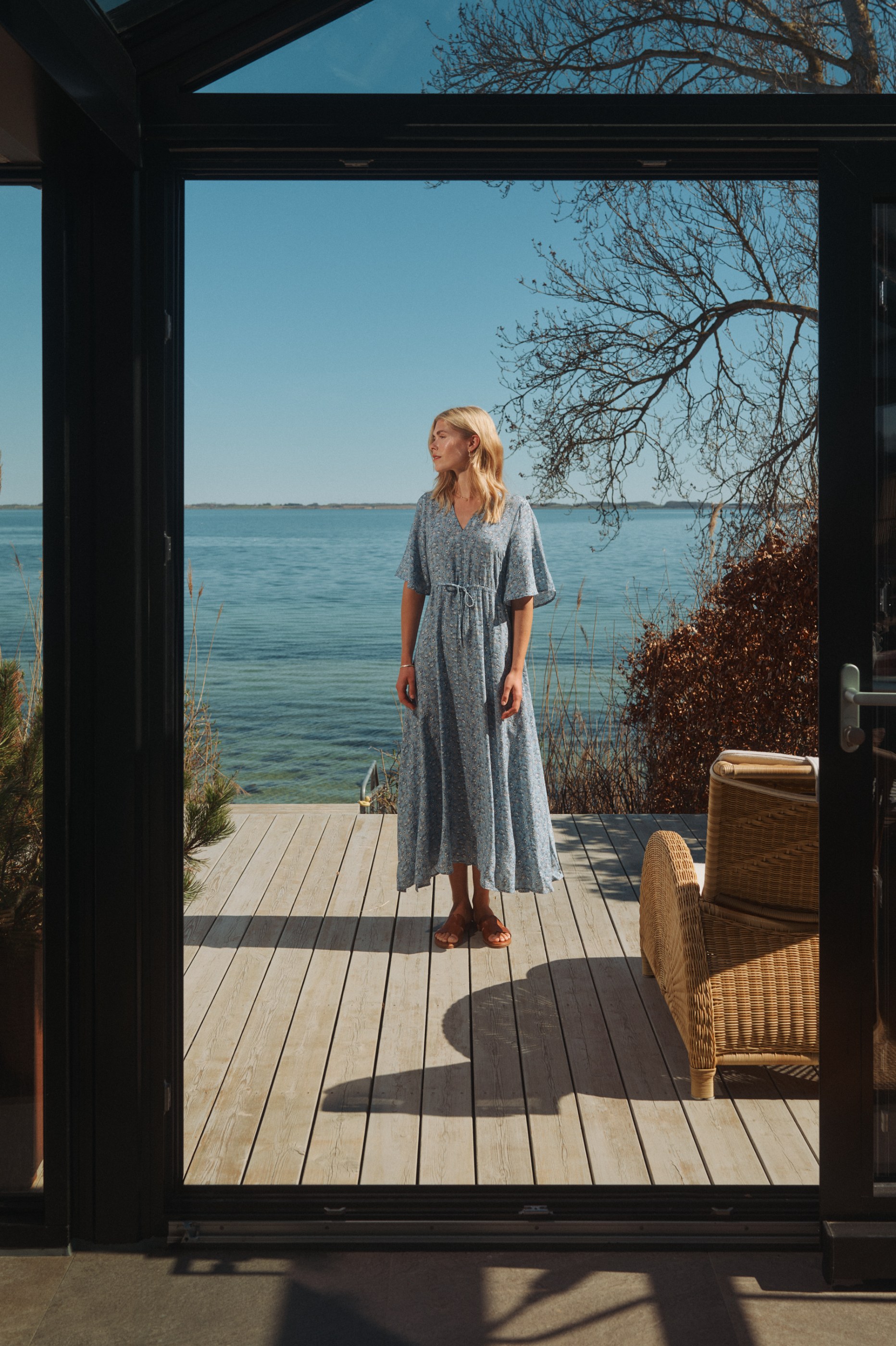Woman in blue floral dress on a sunlit wooden deck overlooking water, framed by a dark doorway.