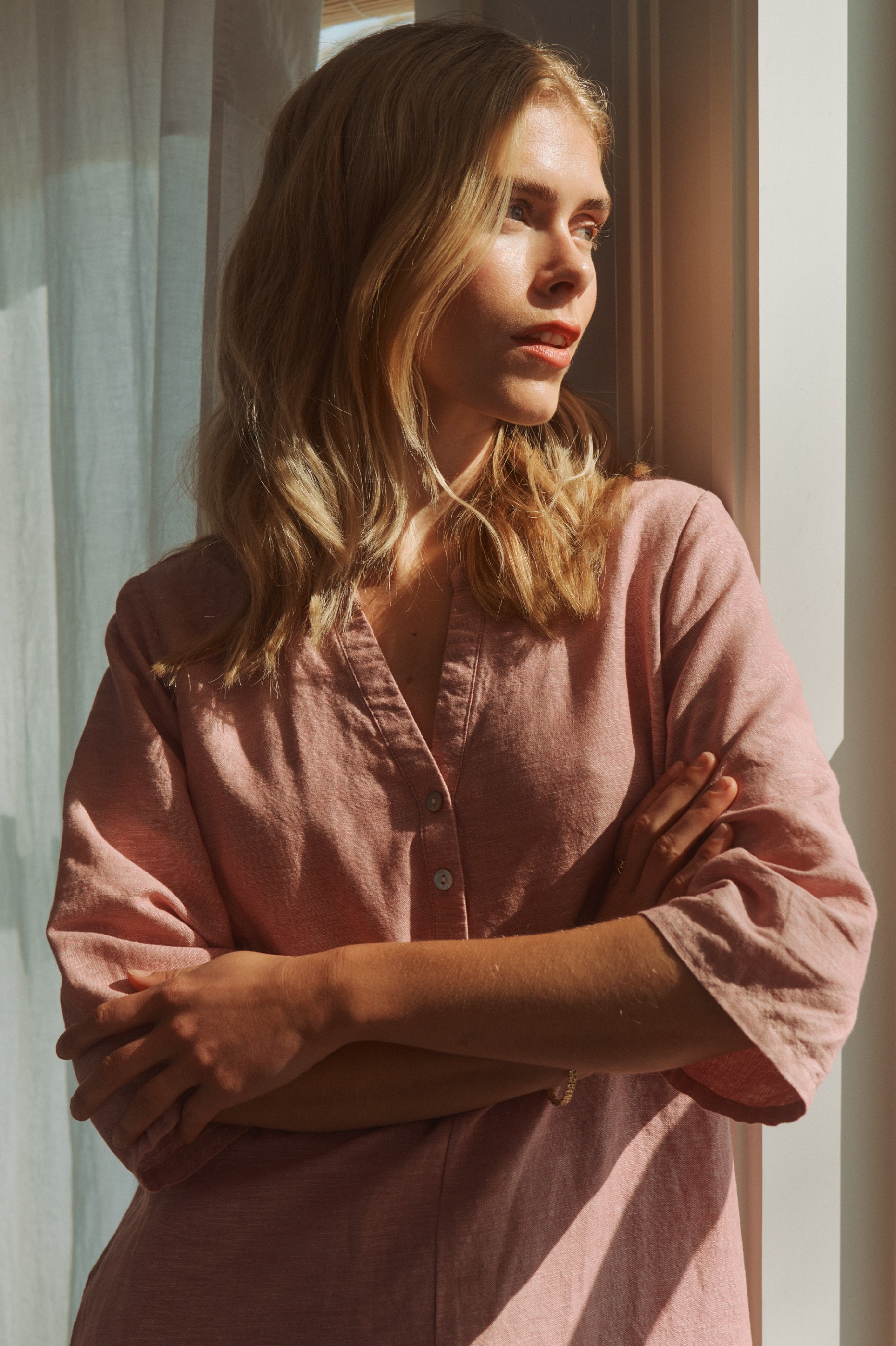 Young woman with blonde hair in a pink linen shirt, arms crossed, looking out a sunlit window.