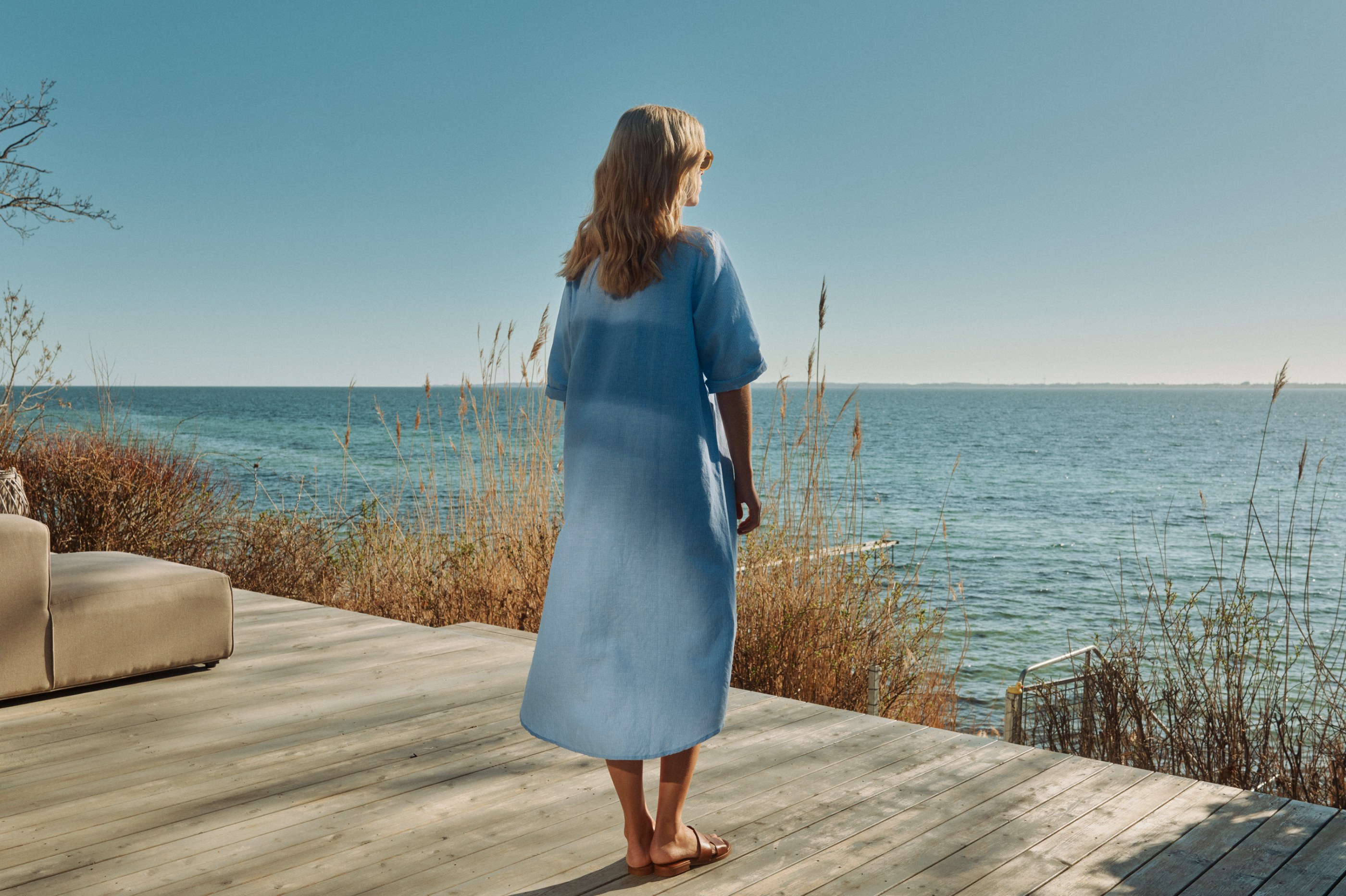 Blonde woman in a light blue dress stands on a wooden deck overlooking the sea.