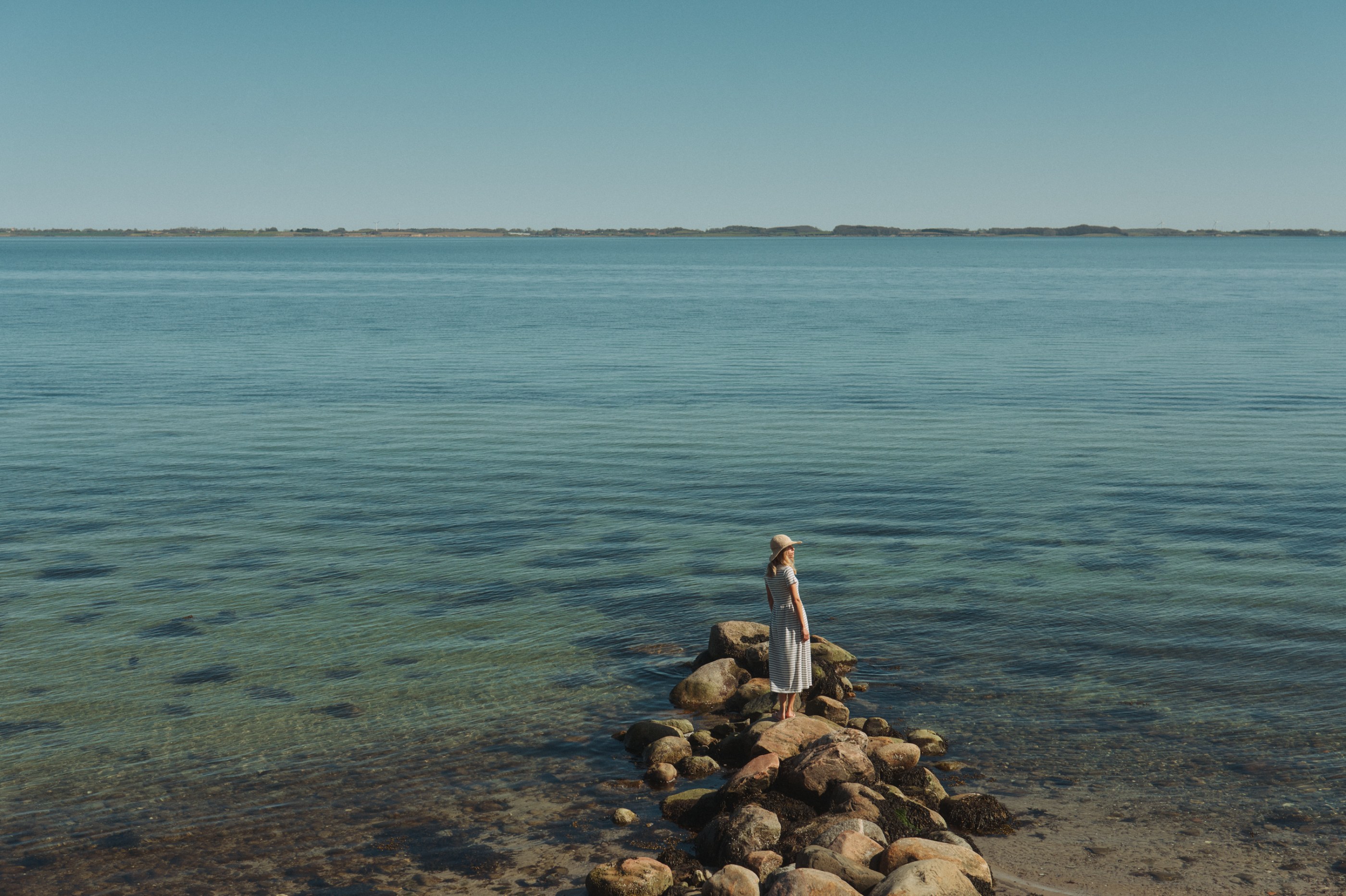 Woman in striped dress and hat on rocks, looking at clear sea and distant land.