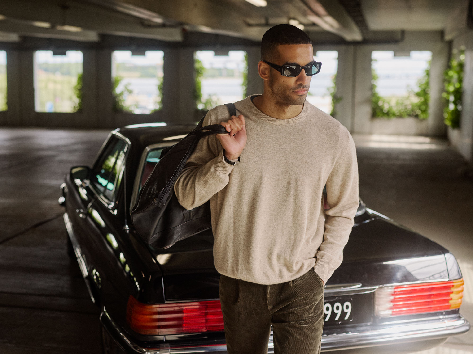 Man in a beige sweater and dark sunglasses carrying a bag by a black vintage car.