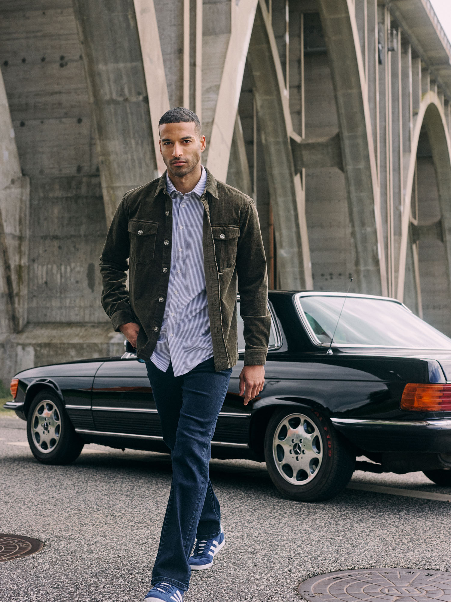 Man in green corduroy jacket and jeans walks by a vintage black car and bridge.