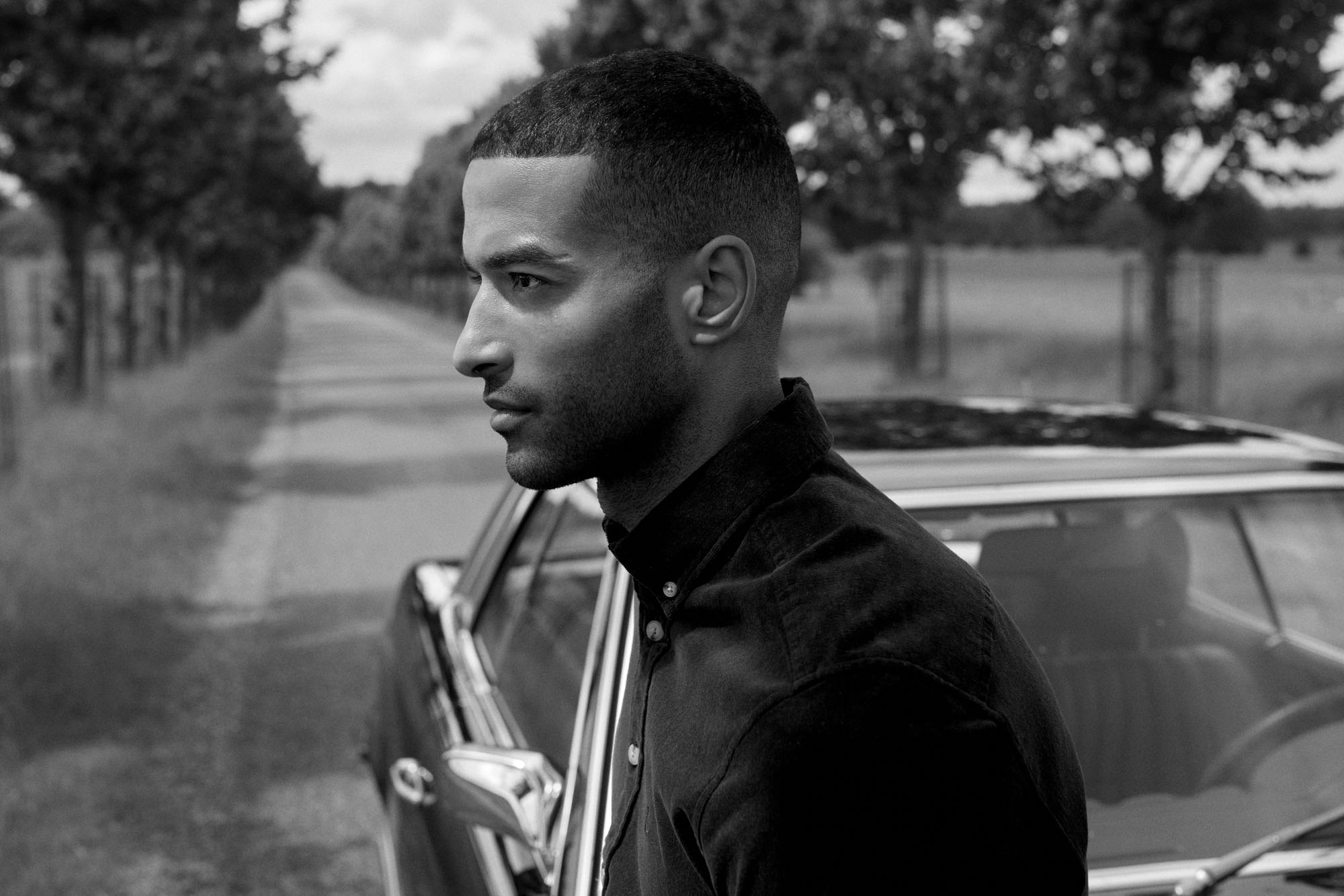 Monochrome profile of a man in dark shirt by classic car on tree-lined road.