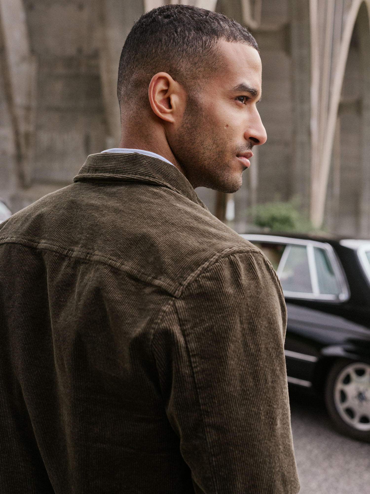 Man in olive corduroy jacket looking over his shoulder with urban arches and a car in the background.