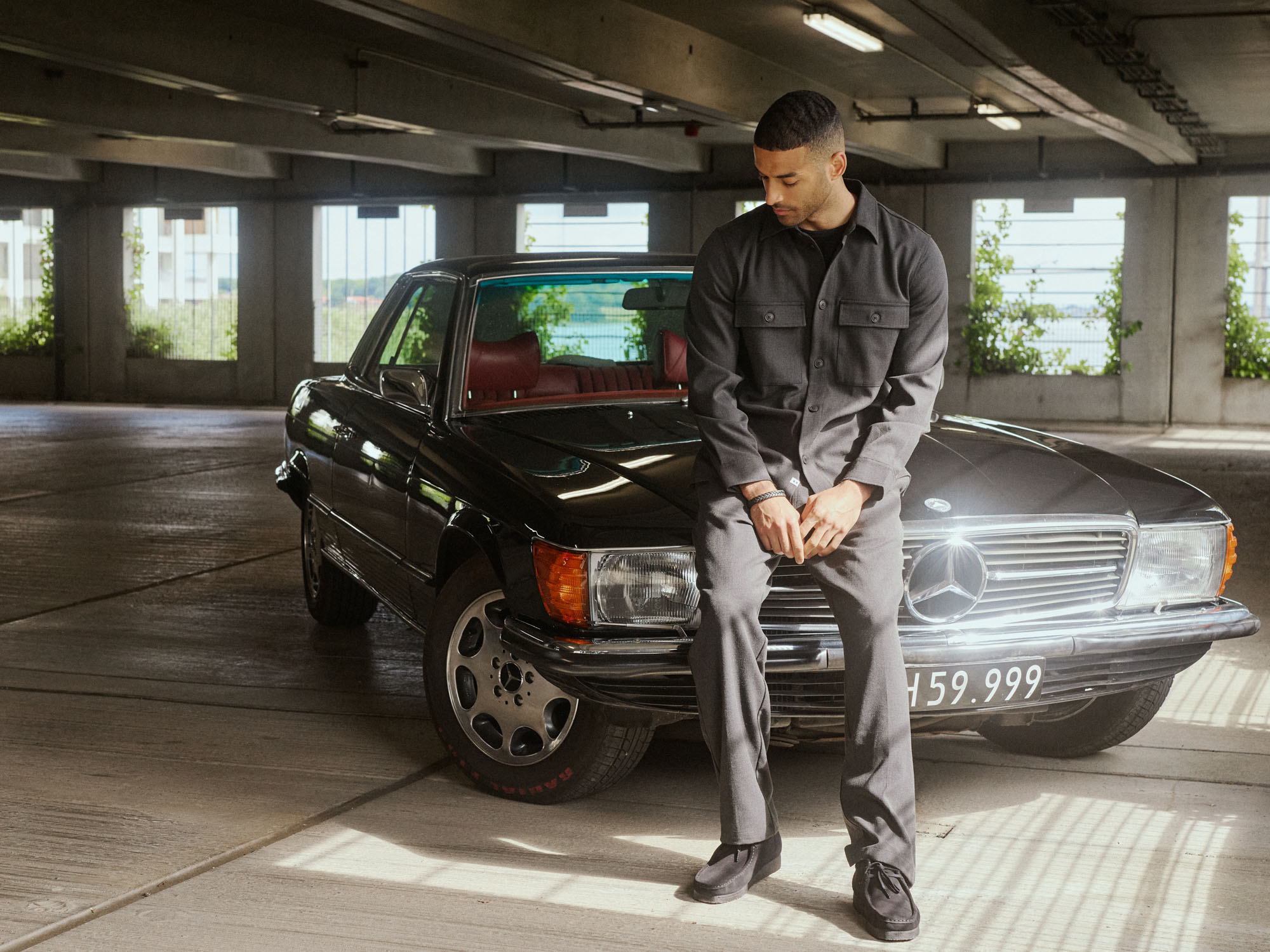 Man in grey shirt sits on black classic car in a sunlit parking garage.