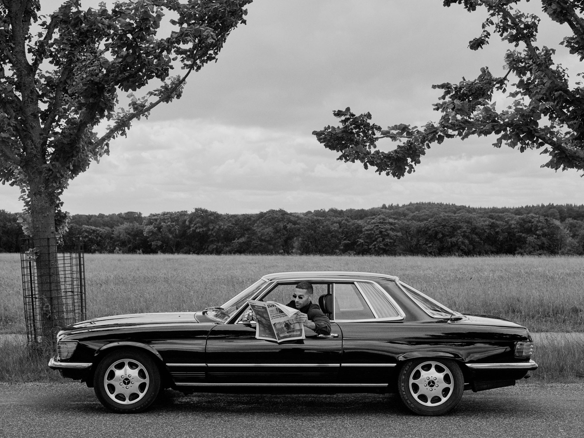 Man in sunglasses reading a newspaper in a black coupe on a rural road.