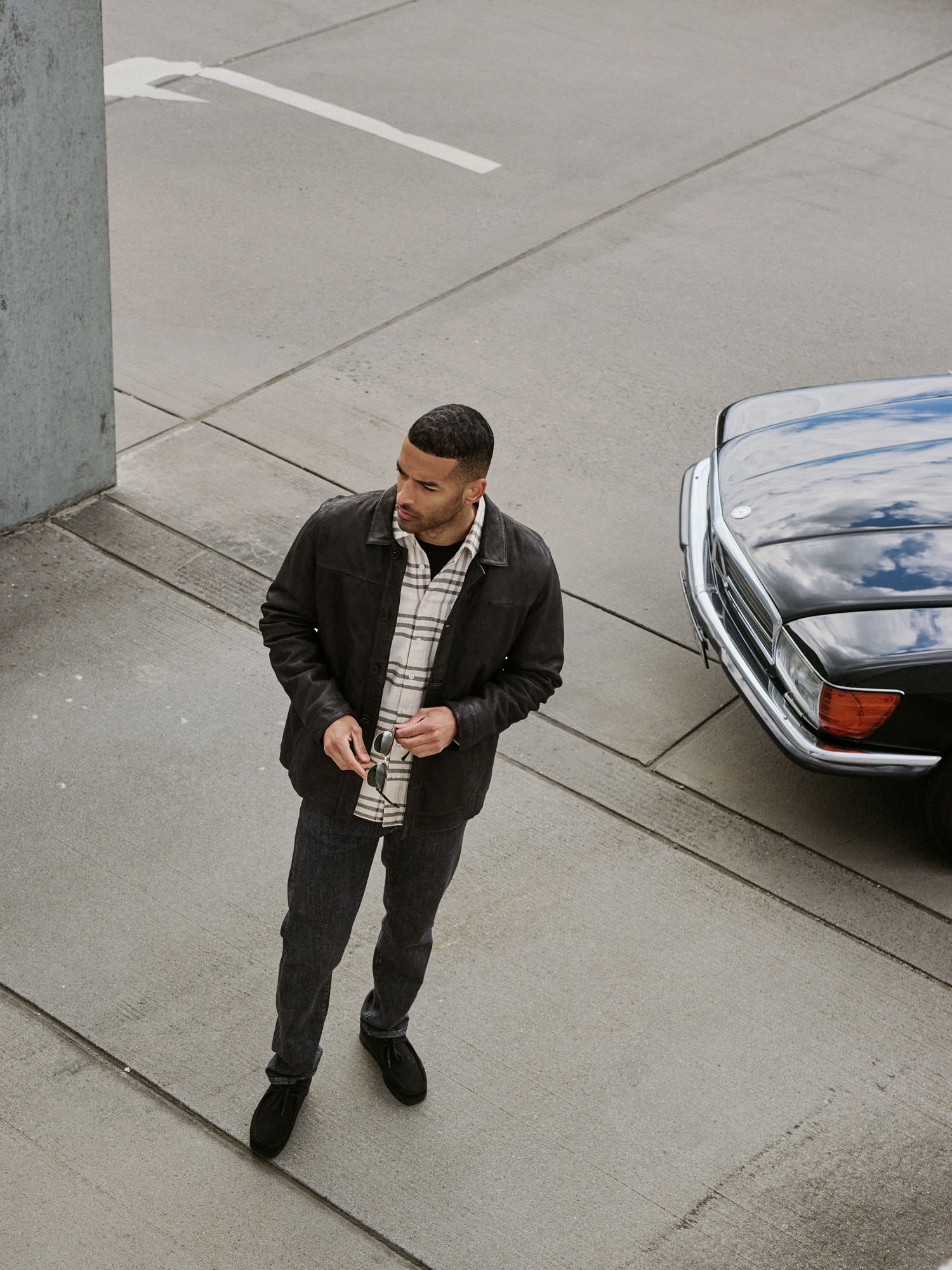 Man in dark jacket, plaid shirt, holding sunglasses, standing next to a black car.