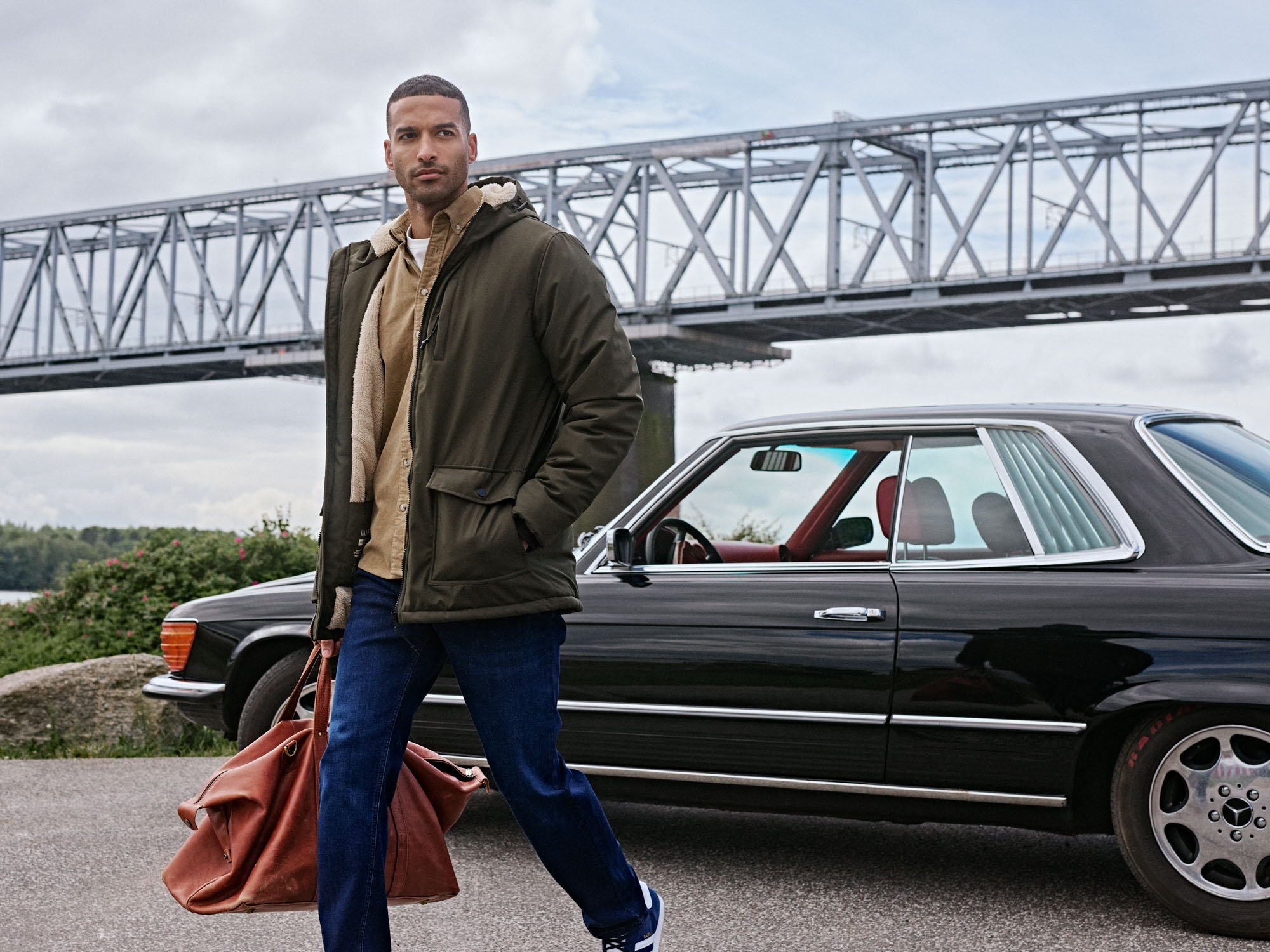 Man in green jacket and jeans with a leather bag by a classic car and bridge.