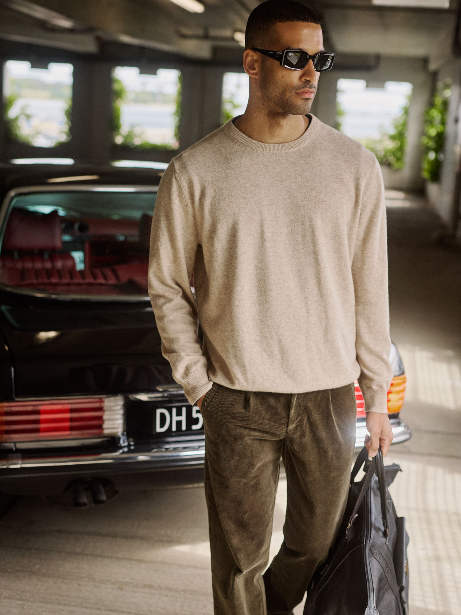Man wearing a beige sweater, olive corduroy pants, sunglasses, by a classic car.