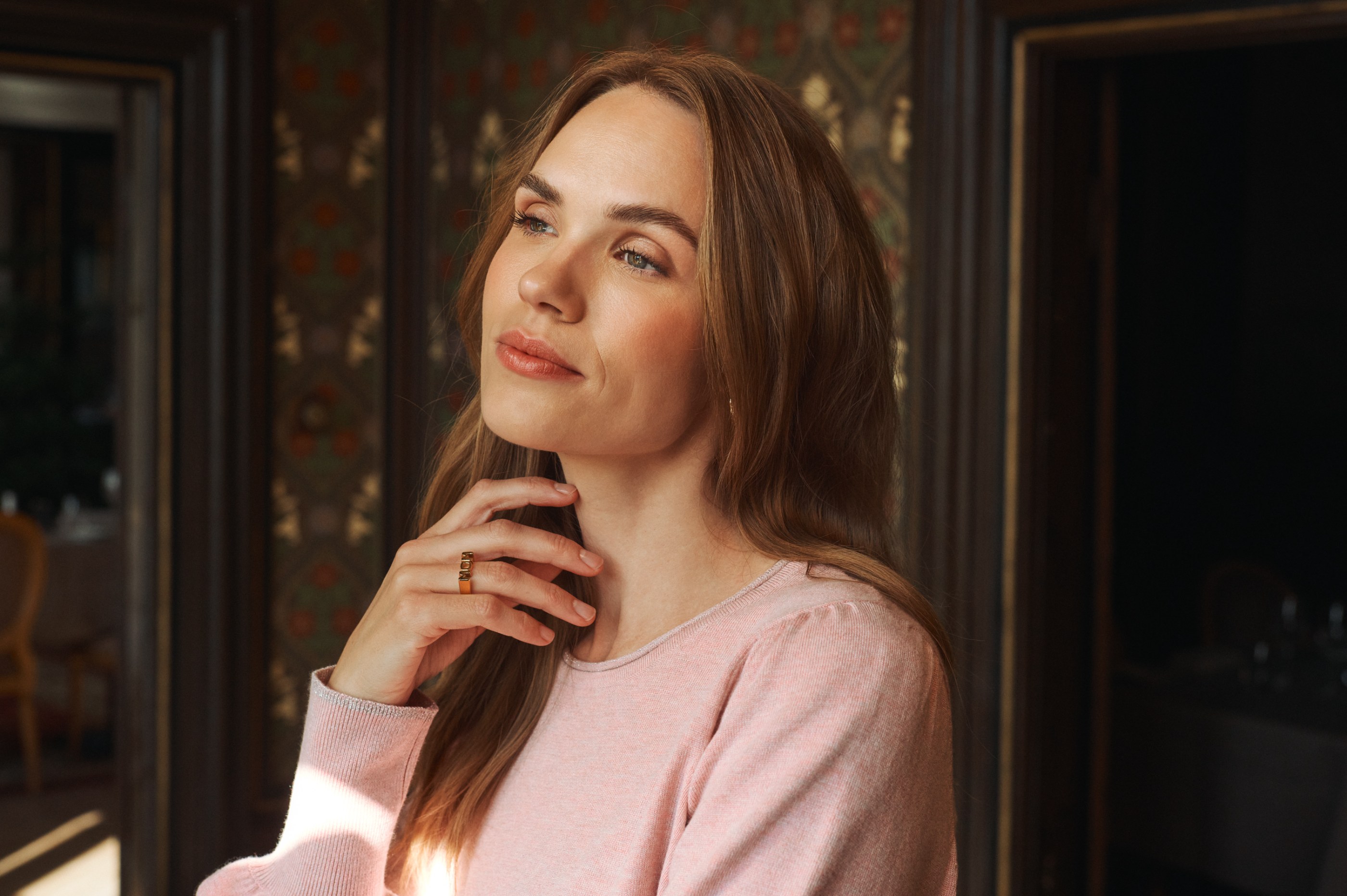 Woman in a pink sweater with a 'MOM' ring, gazing upward in a warmly lit room.