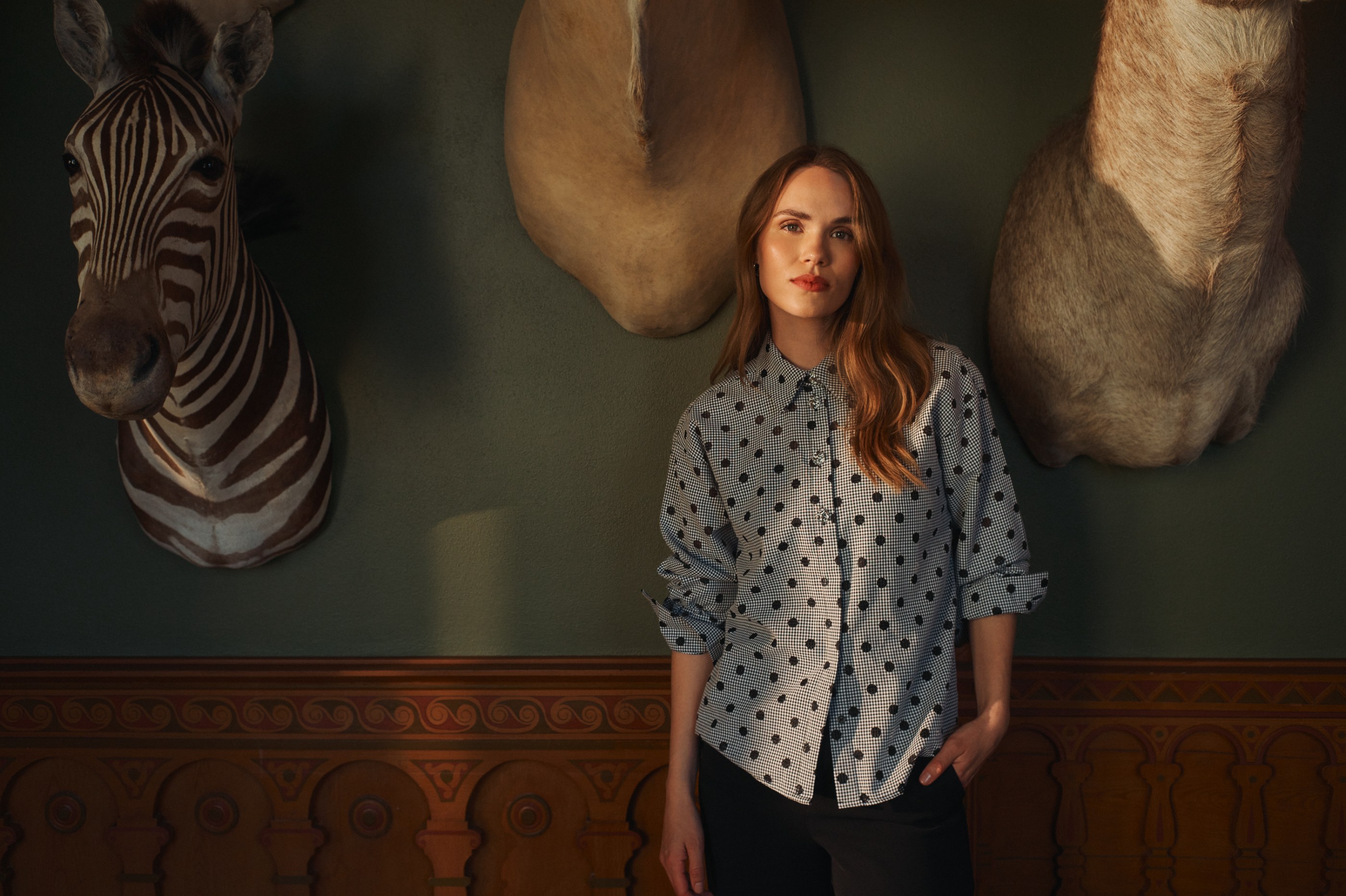 Woman in a polka-dot shirt stands under dramatic light, flanked by taxidermy animal heads.