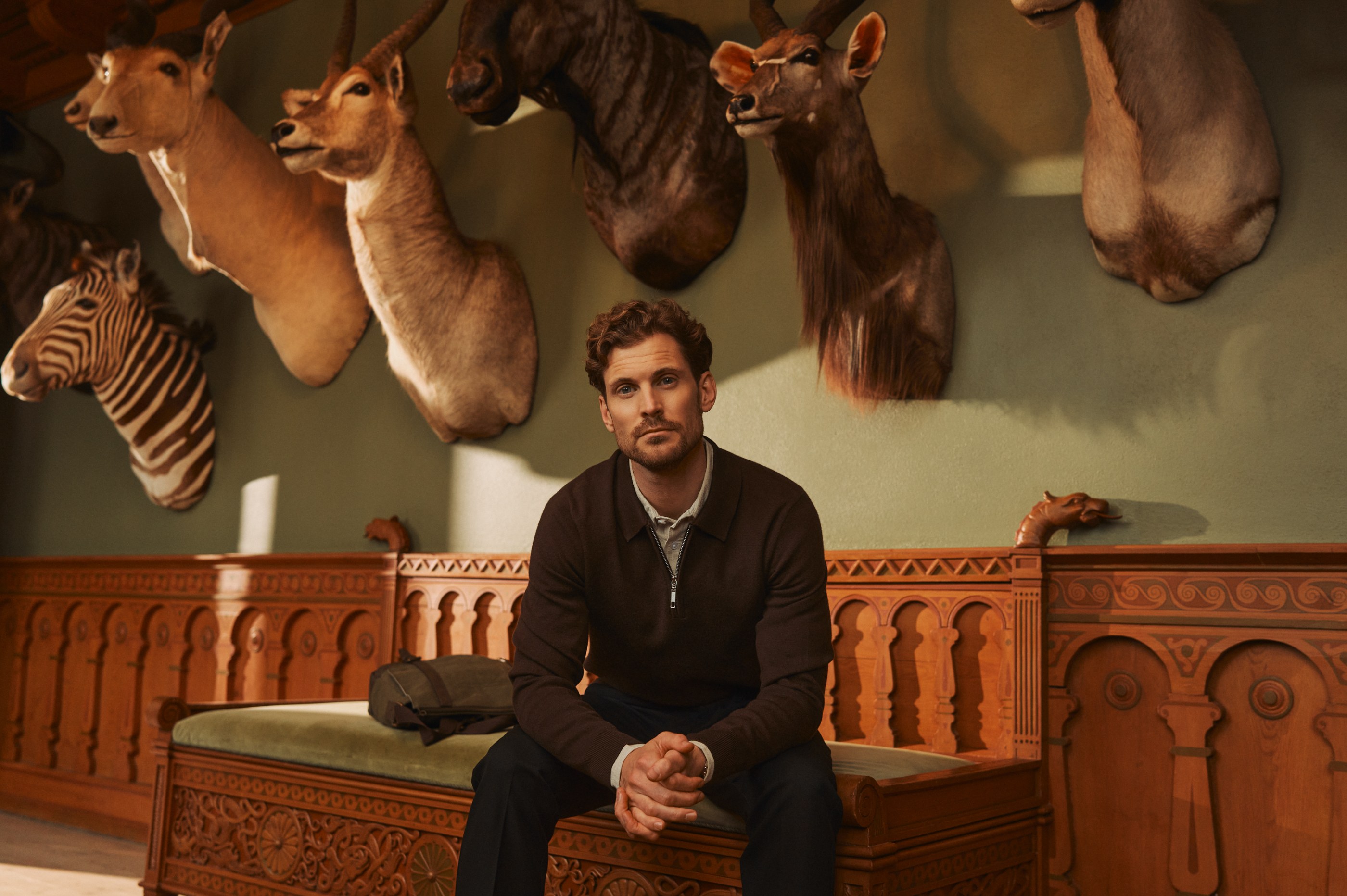 A man in a brown sweater sits on an ornate wooden bench surrounded by taxidermied animal heads.