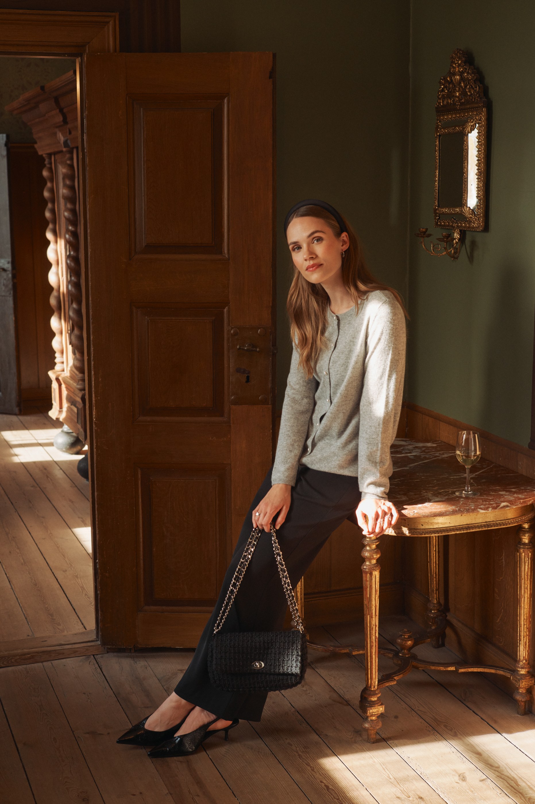 Woman in grey cardigan and black pants holds black bag on sunlit gilded console table.