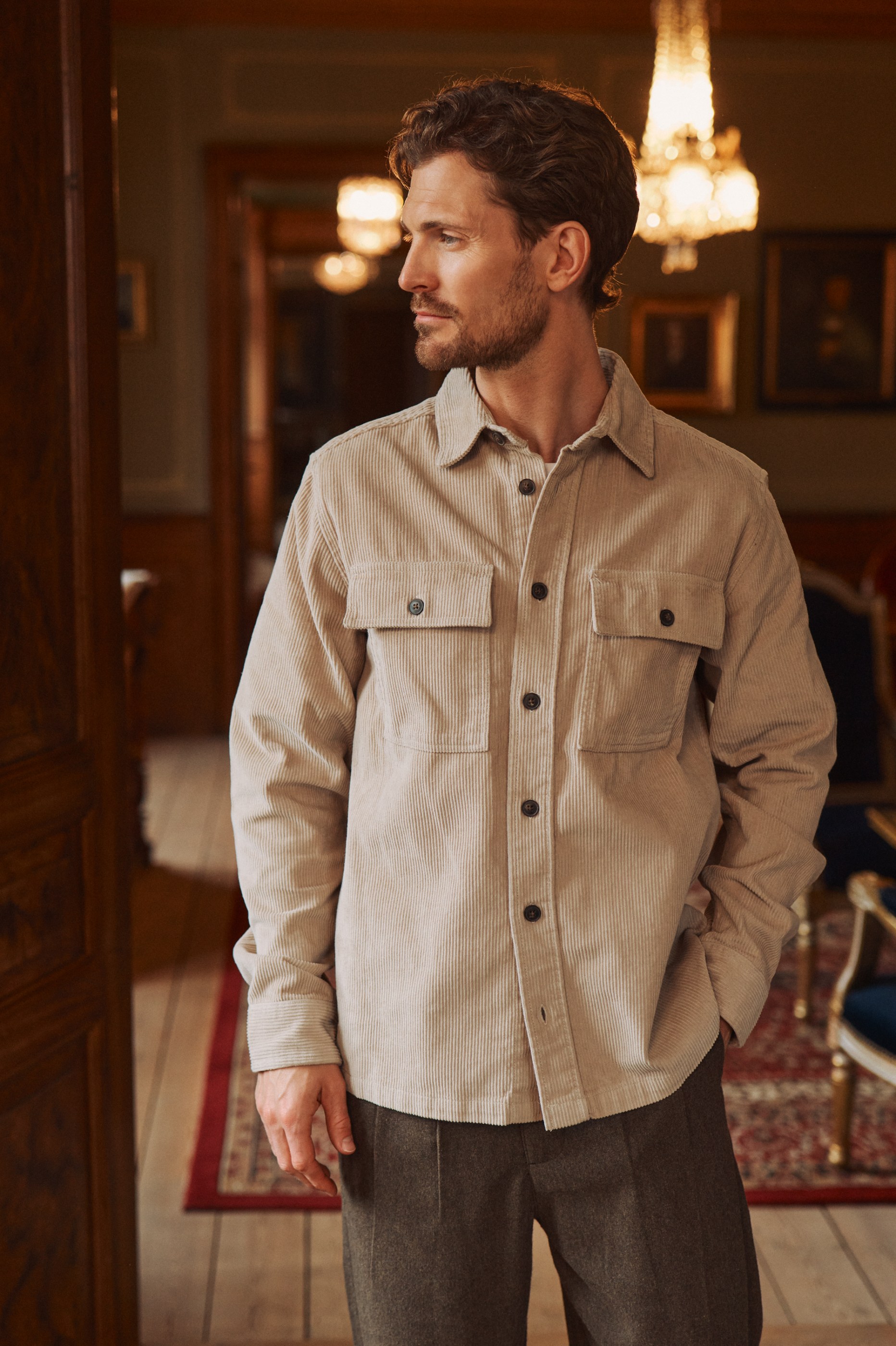 Man in a beige corduroy shirt and dark pants standing in an elegantly lit mansion.