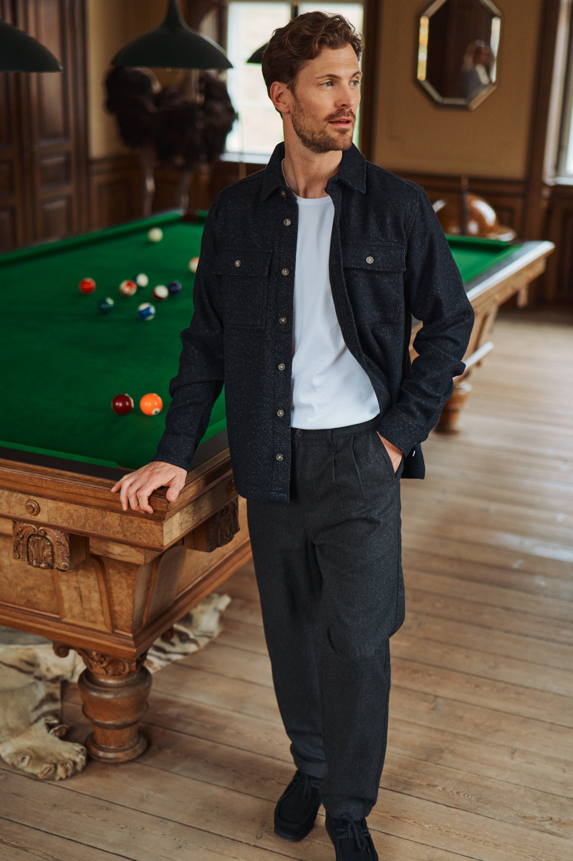 Man in dark shirt and trousers stands by an ornate wooden billiard table in a grand interior.