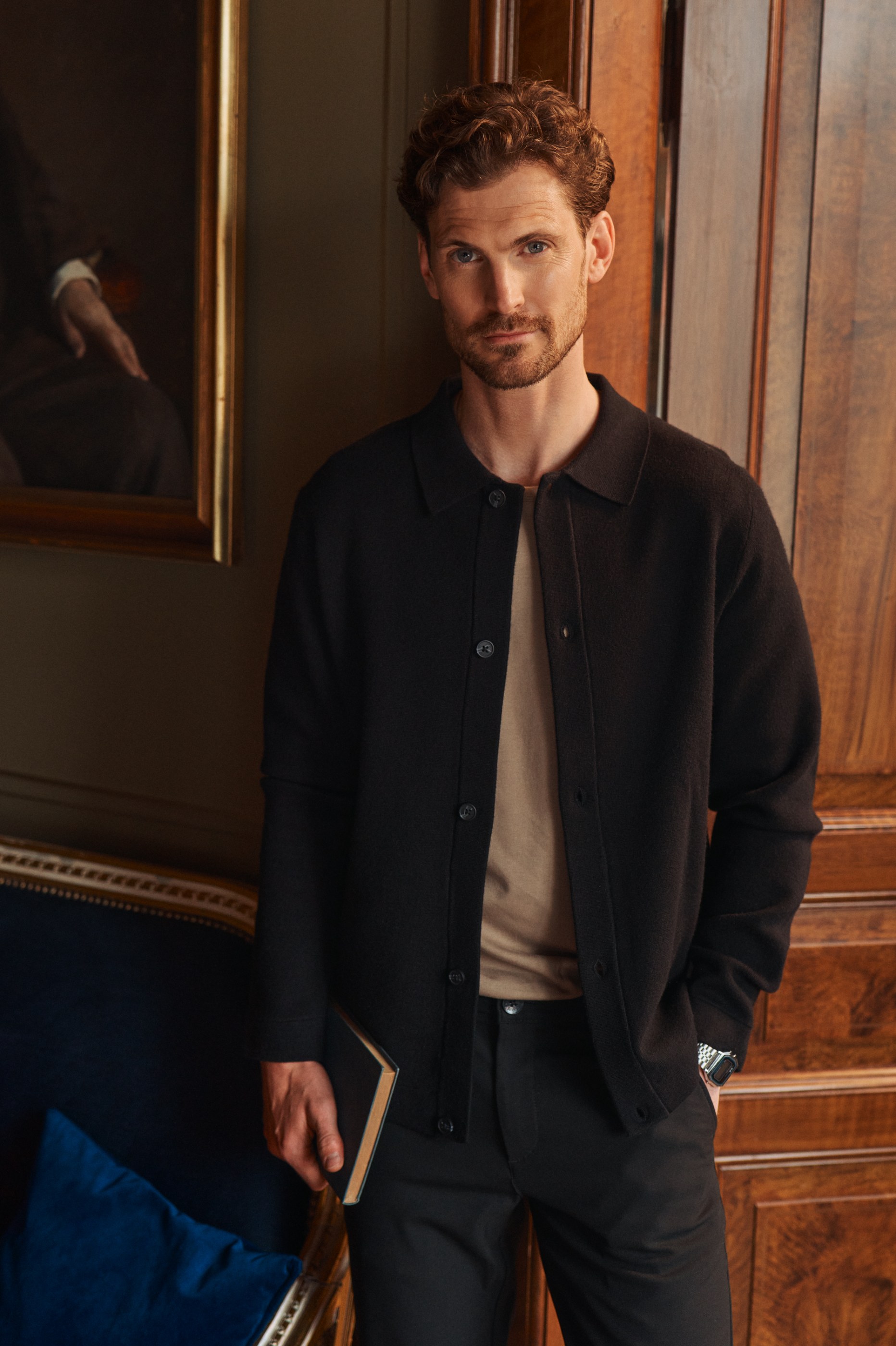 Man in a black cardigan holds a book, standing in an elegant, wood-paneled room.