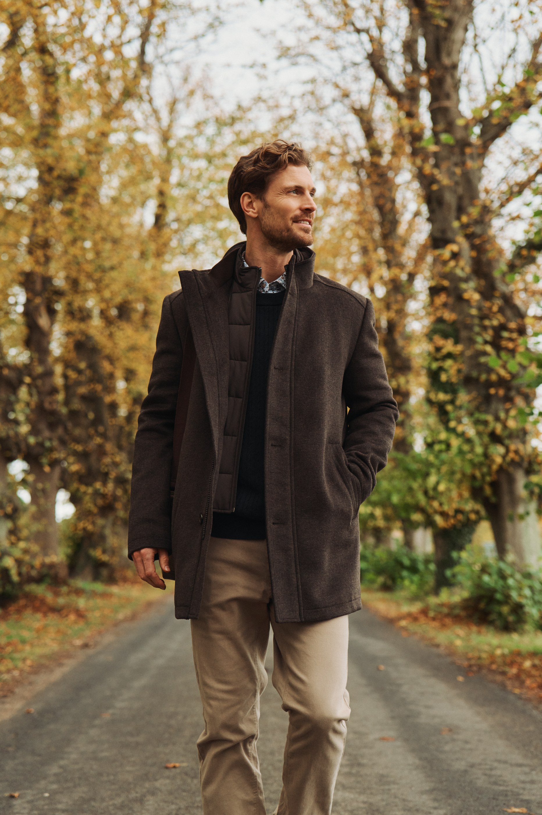 Smiling man in a warm brown coat and khaki pants walks down an autumn tree-lined road.