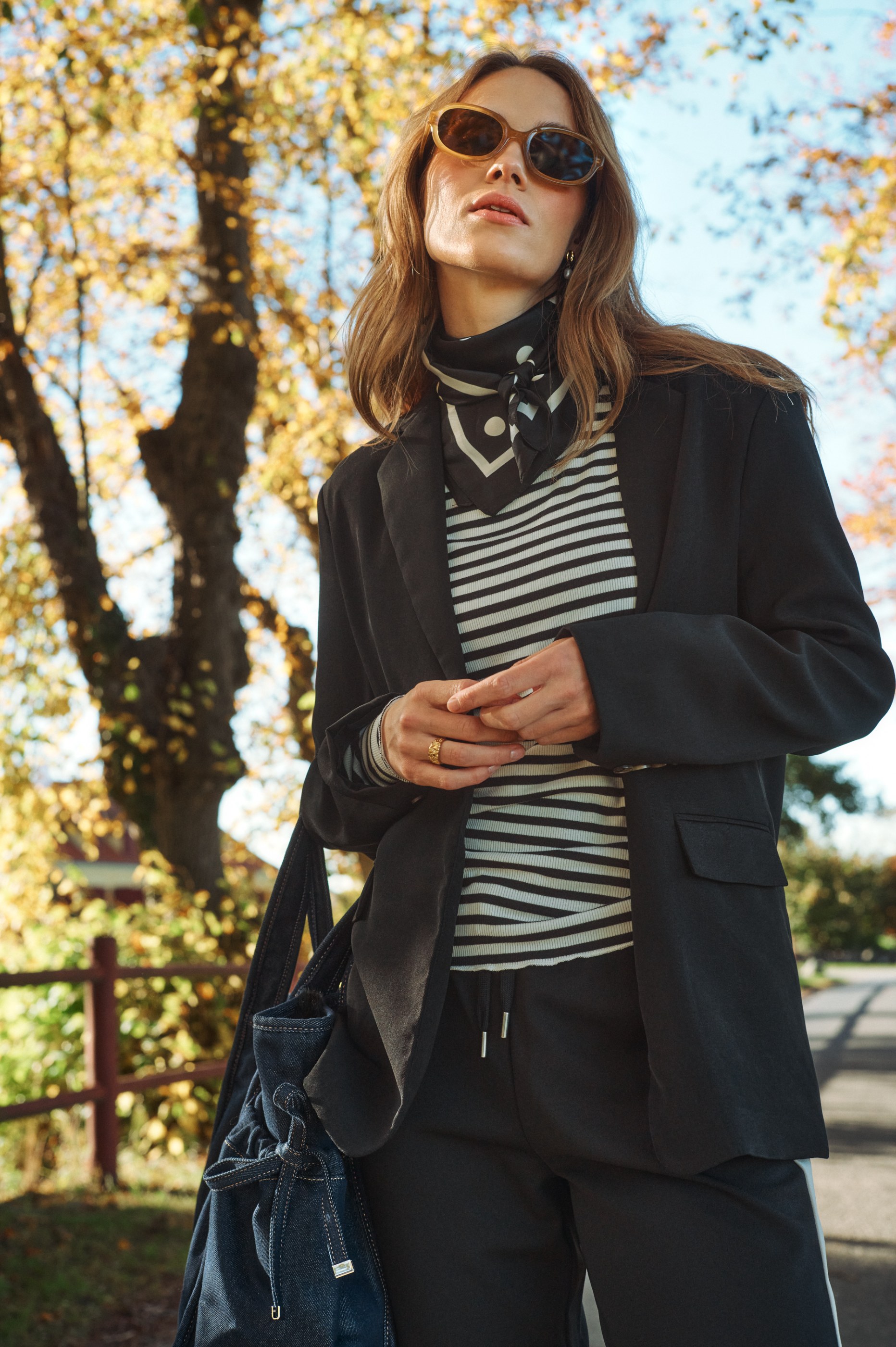 Fashionable woman in black blazer, striped top, and sunglasses stands among golden autumn trees.