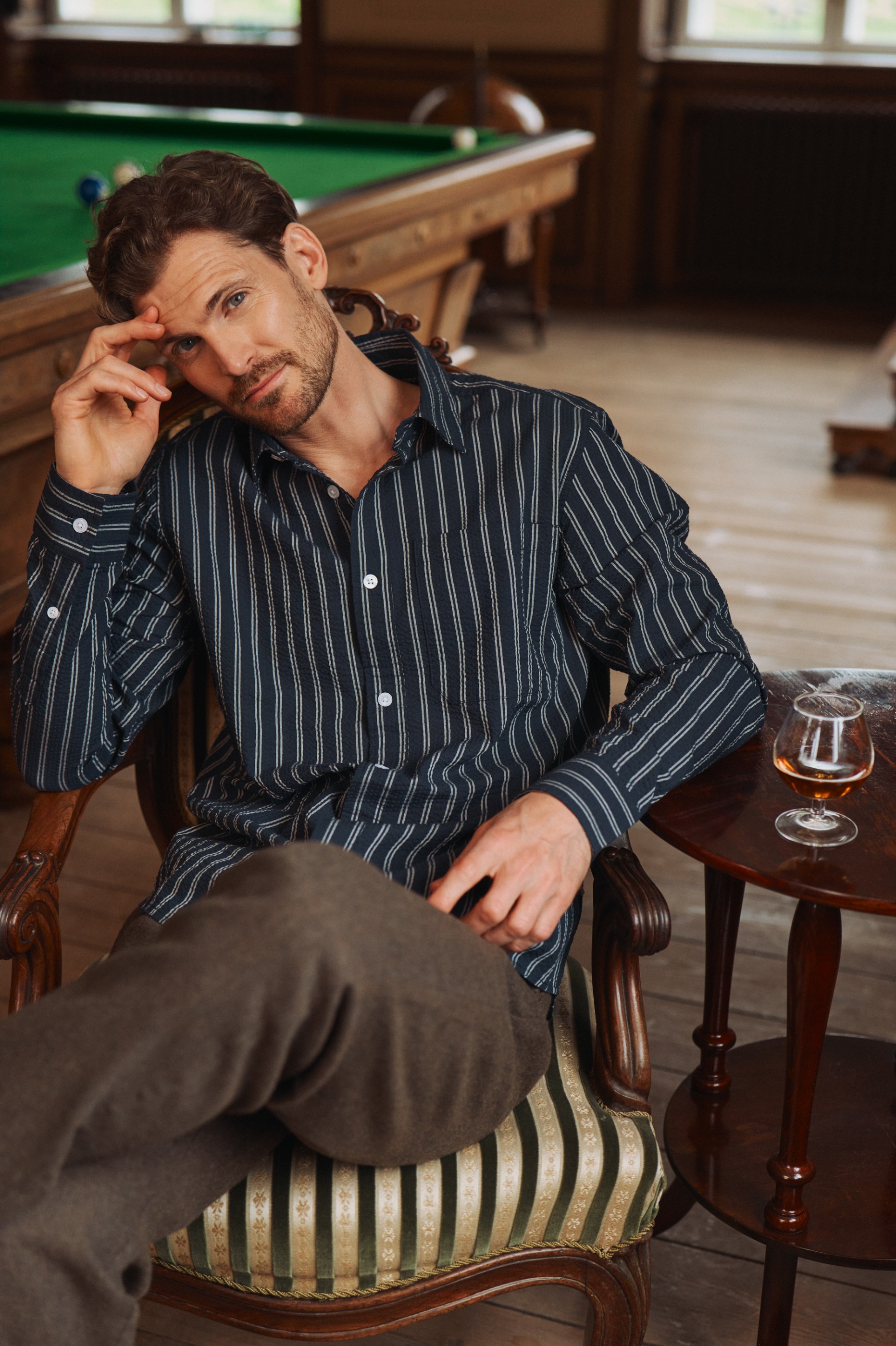 Man in striped shirt sits in ornate chair with drink and pool table.
