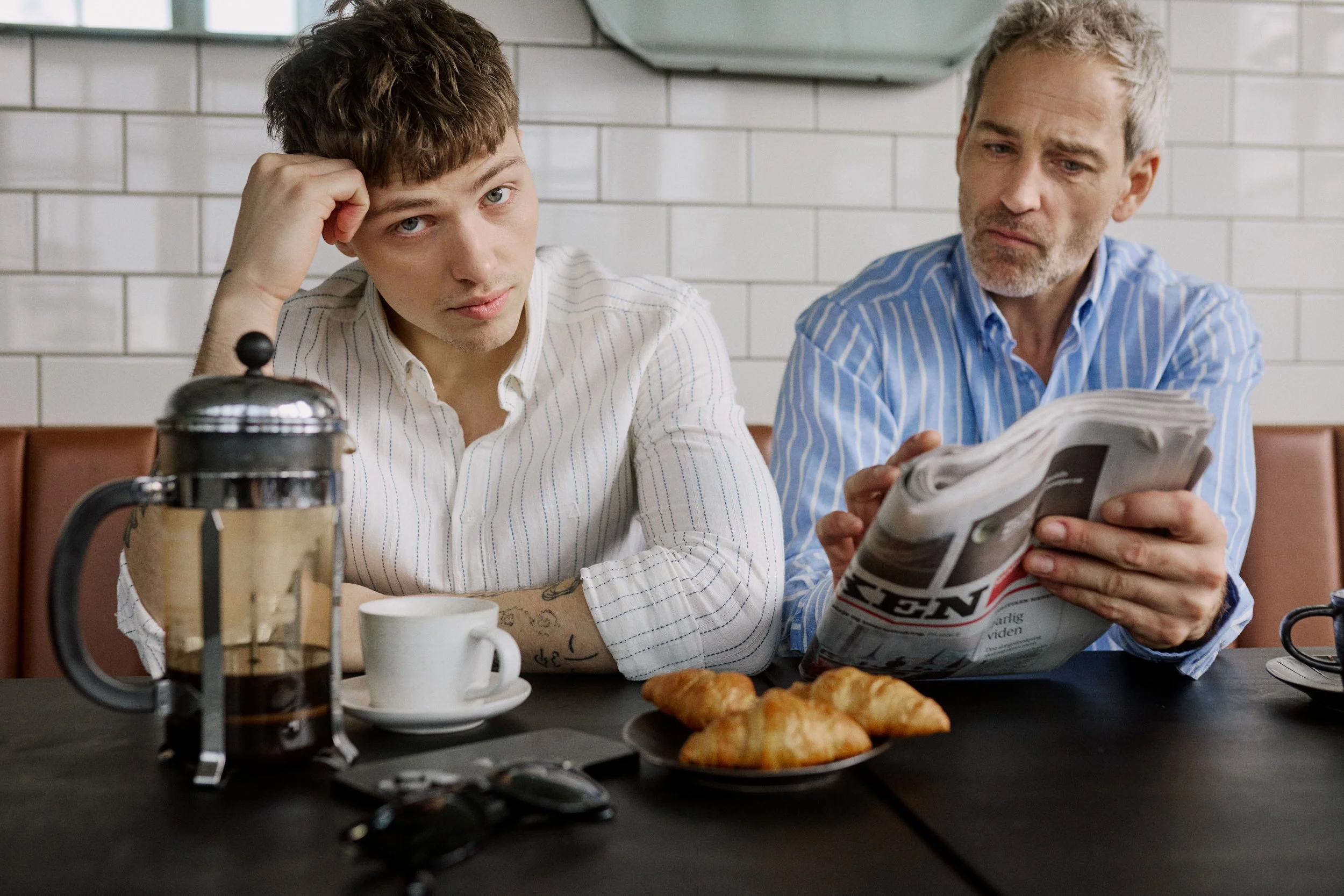 Two men at a cafe table with coffee and croissants, one reading a newspaper.