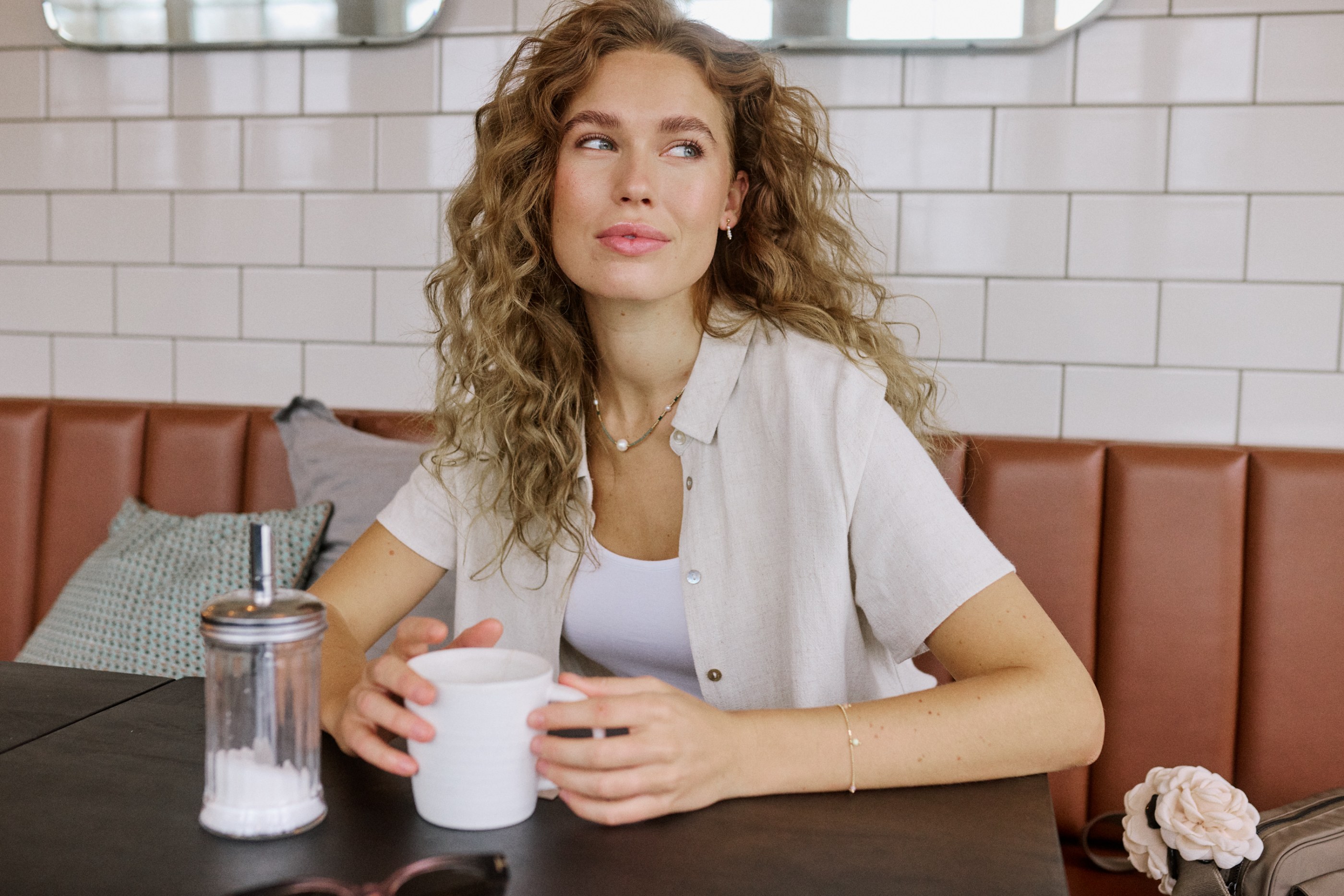 A woman with curly hair sits in a restaurant booth, holding a white mug and looking away.