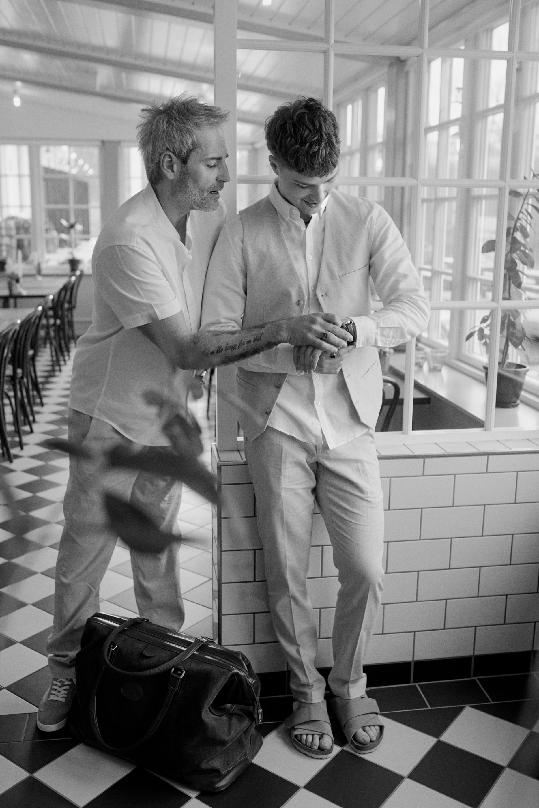 Two men in light casual outfits checking a watch in a bright cafe with a checkerboard floor.