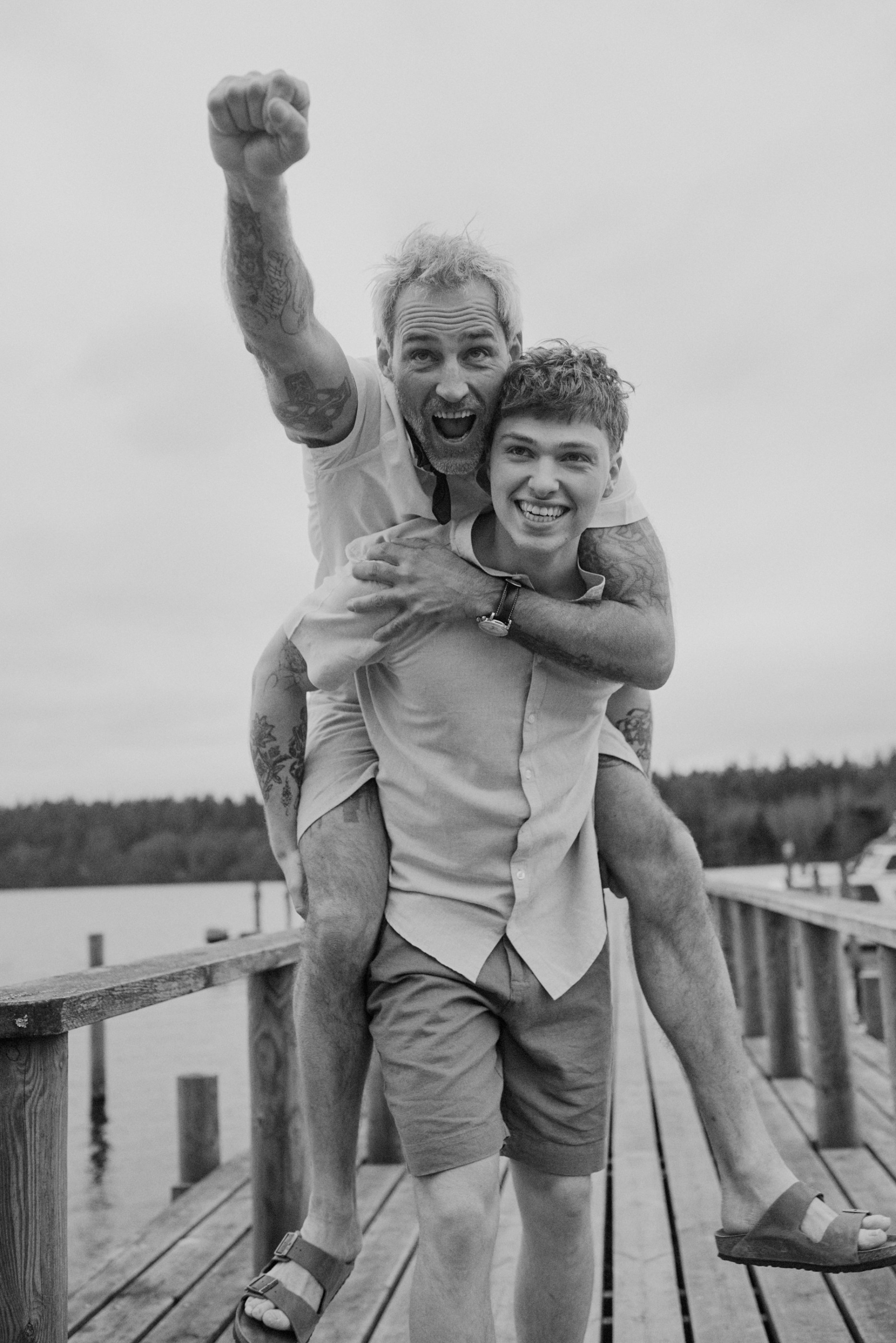 Two smiling men on a wooden pier, one giving a piggyback ride with a raised fist, in black and white.