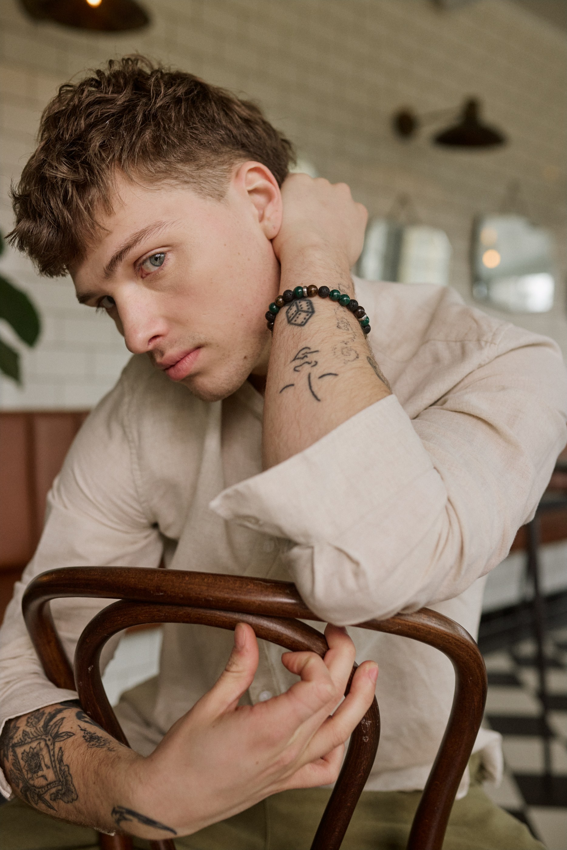 Man with curly brown hair, blue eyes, and arm tattoos leaning on a wooden chair.