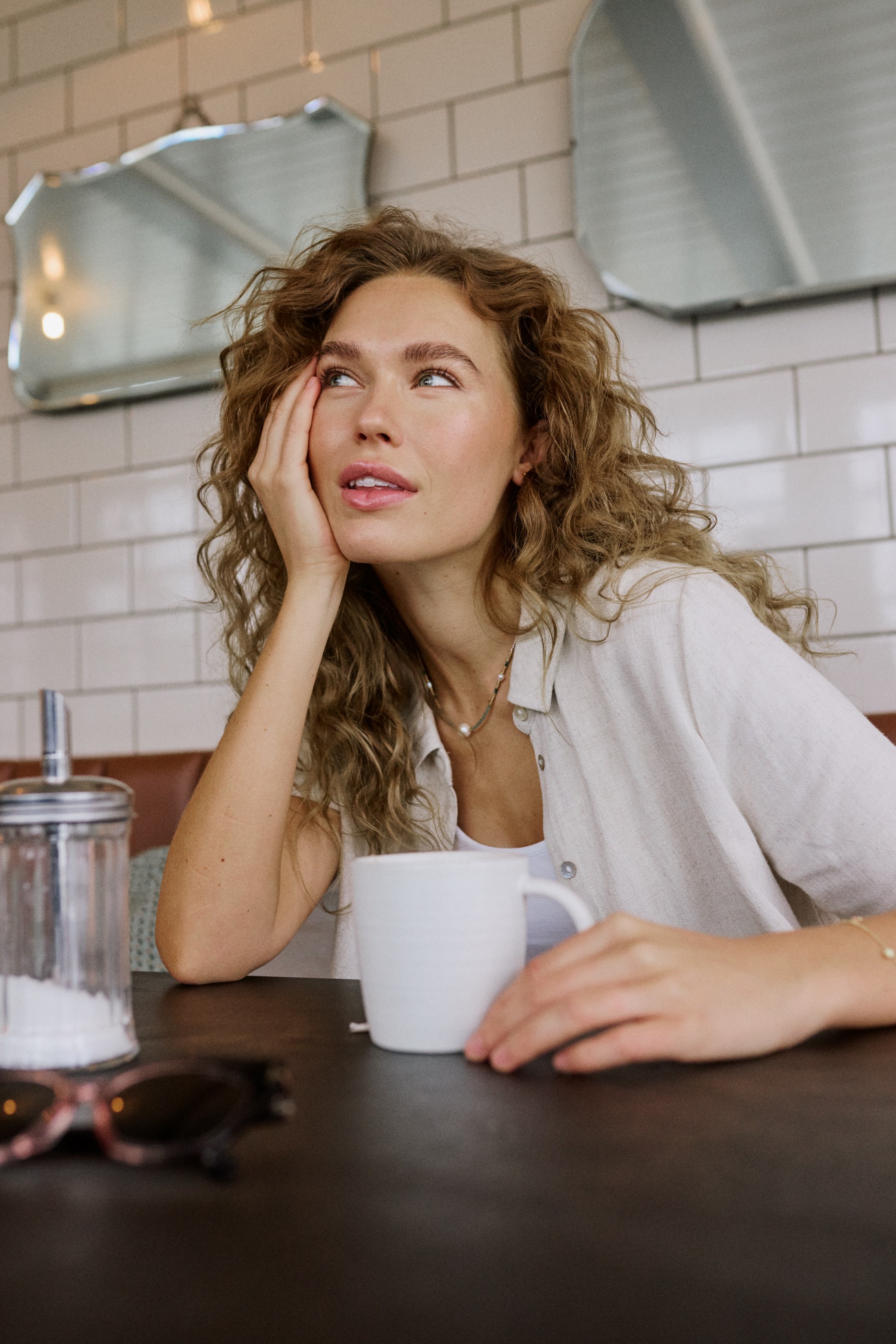 Pensive curly-haired woman looking up at a cafe table with a coffee mug.