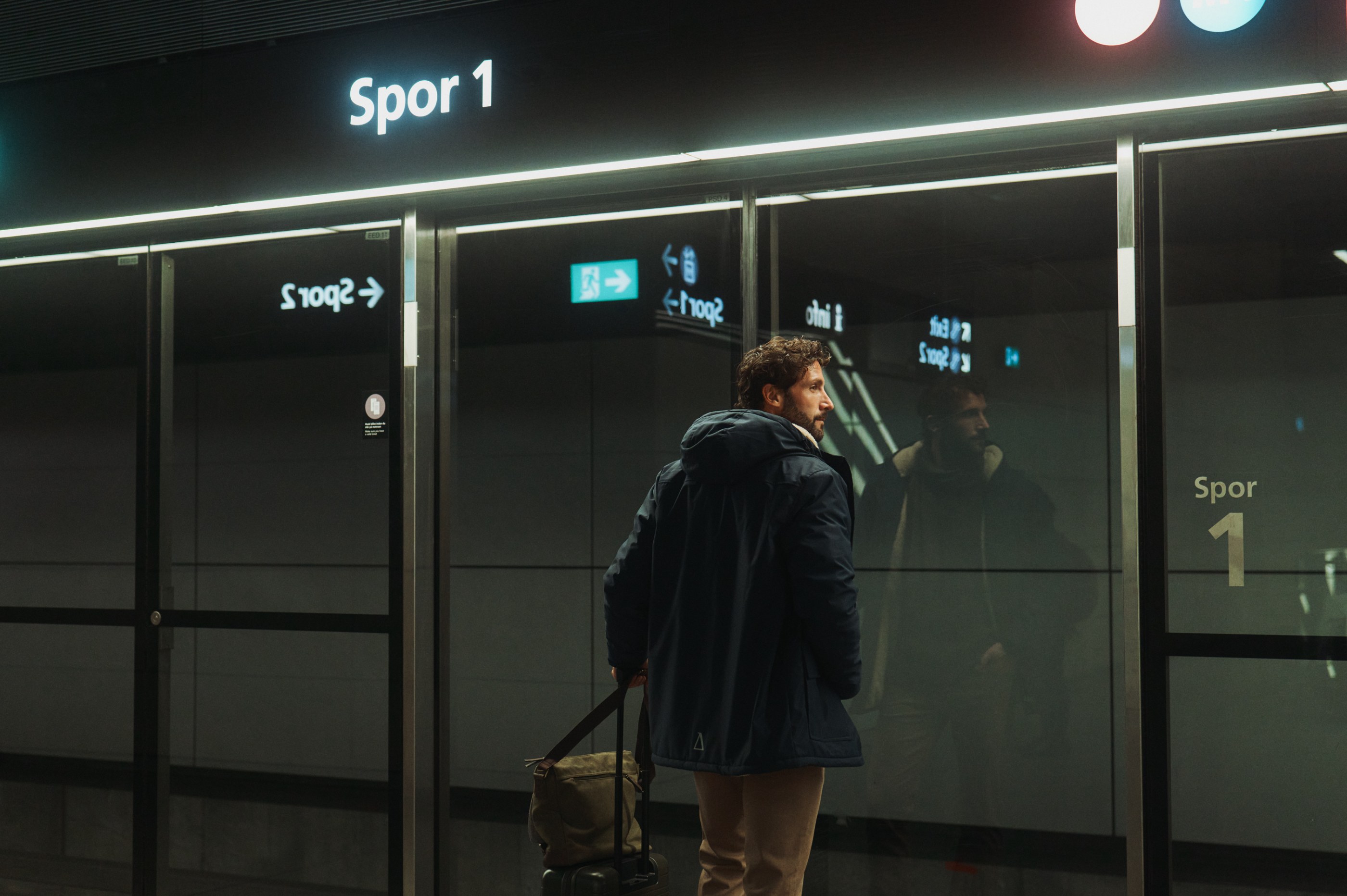 Bearded man in a blue jacket with luggage stands at a modern station with a "Spor 1" sign.