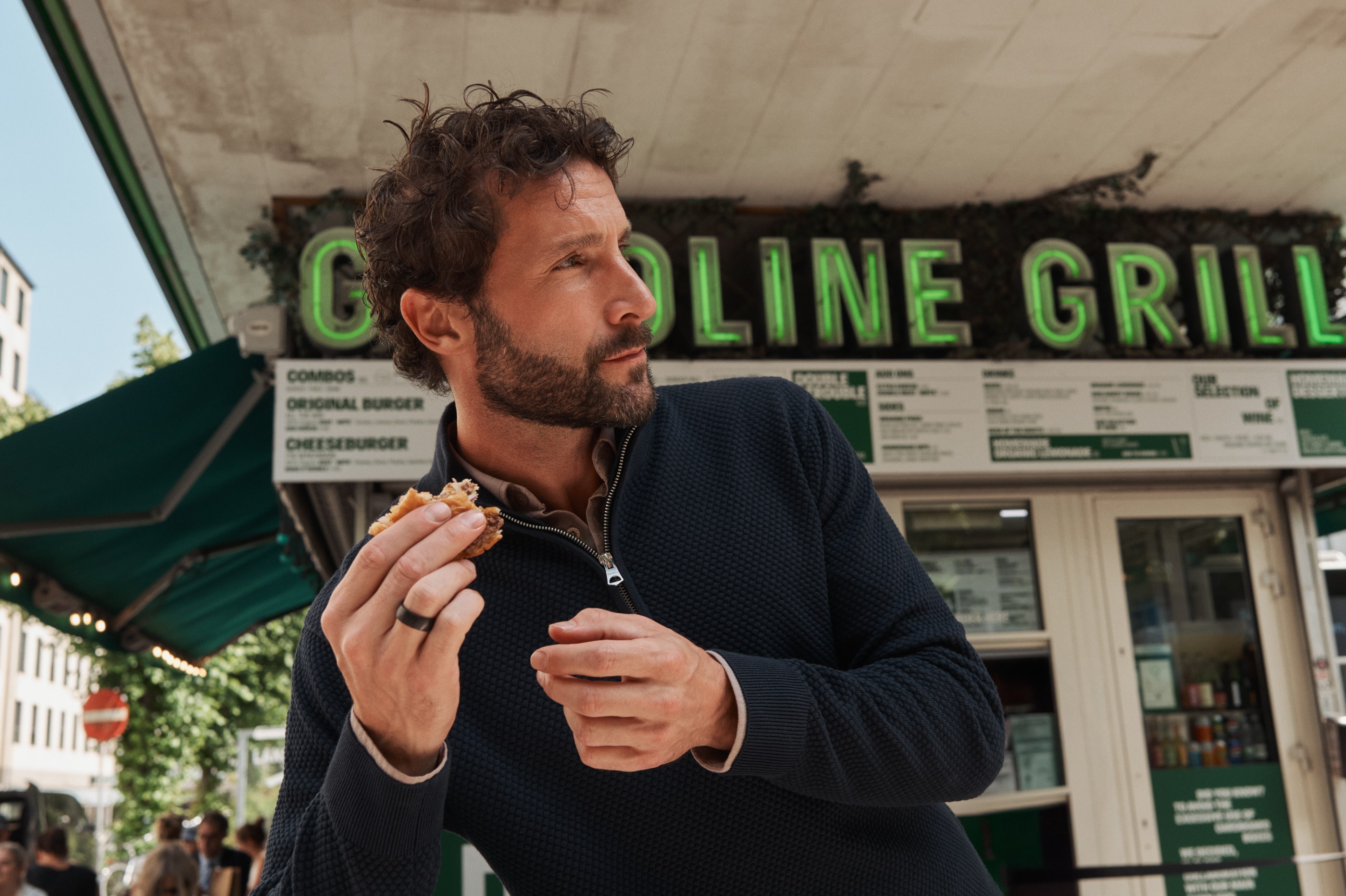 Bearded man in a navy sweater holding a burger at an outdoor Gasoline Grill.