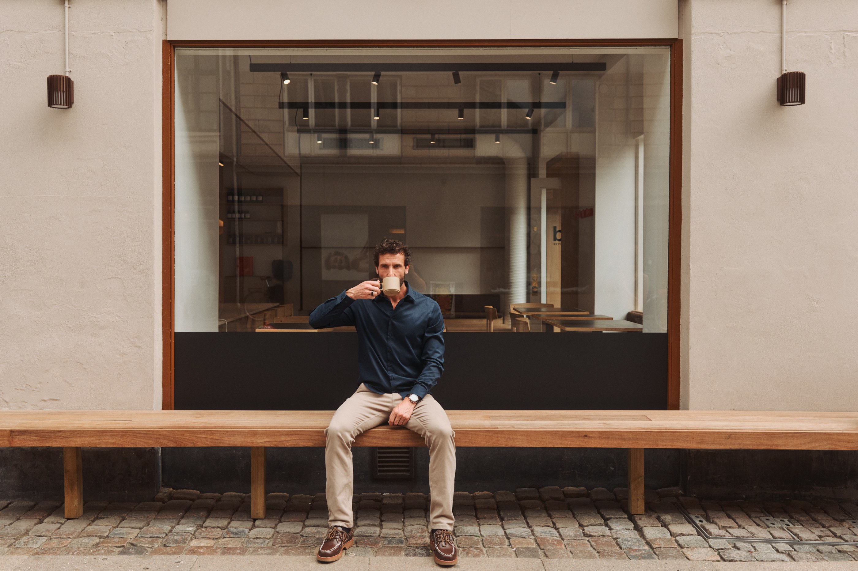 Man in blue shirt sitting on wooden bench drinking from a mug outside a modern cafe.