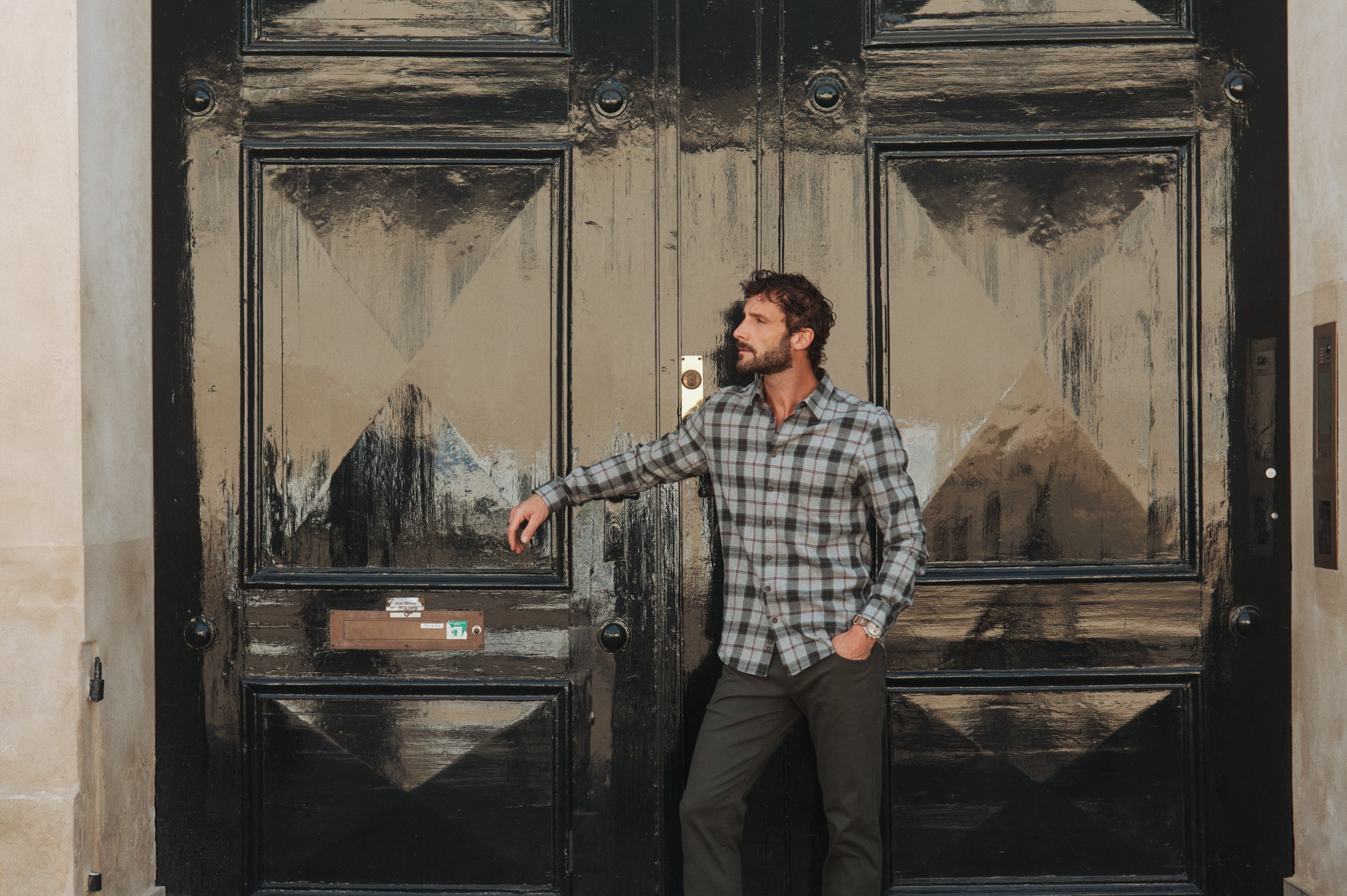 Bearded man in plaid shirt and dark pants leans on a glossy black door.