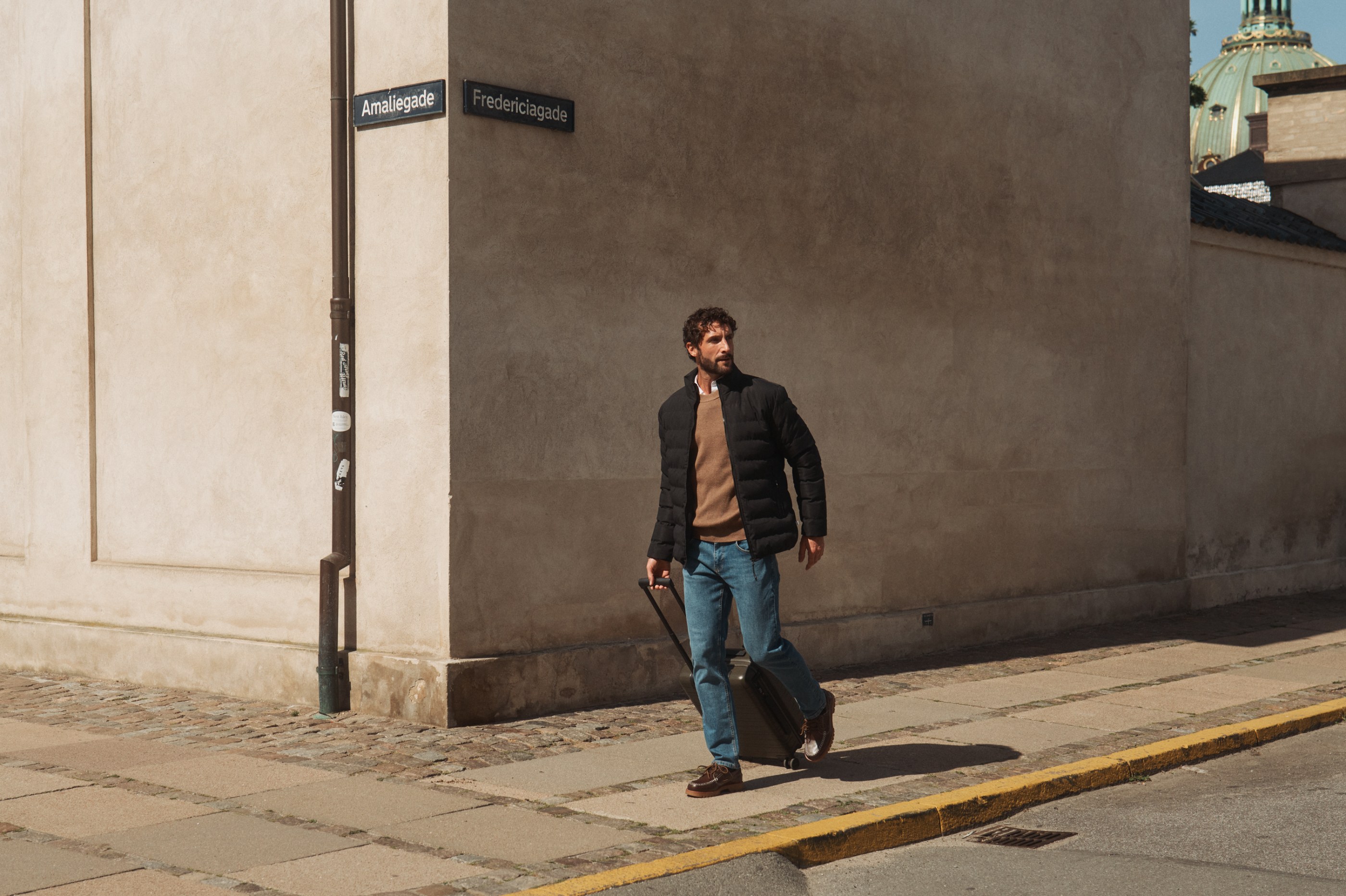 Man in black puffer jacket pulls suitcase on sunny Amaliegade Fredericiagade street.