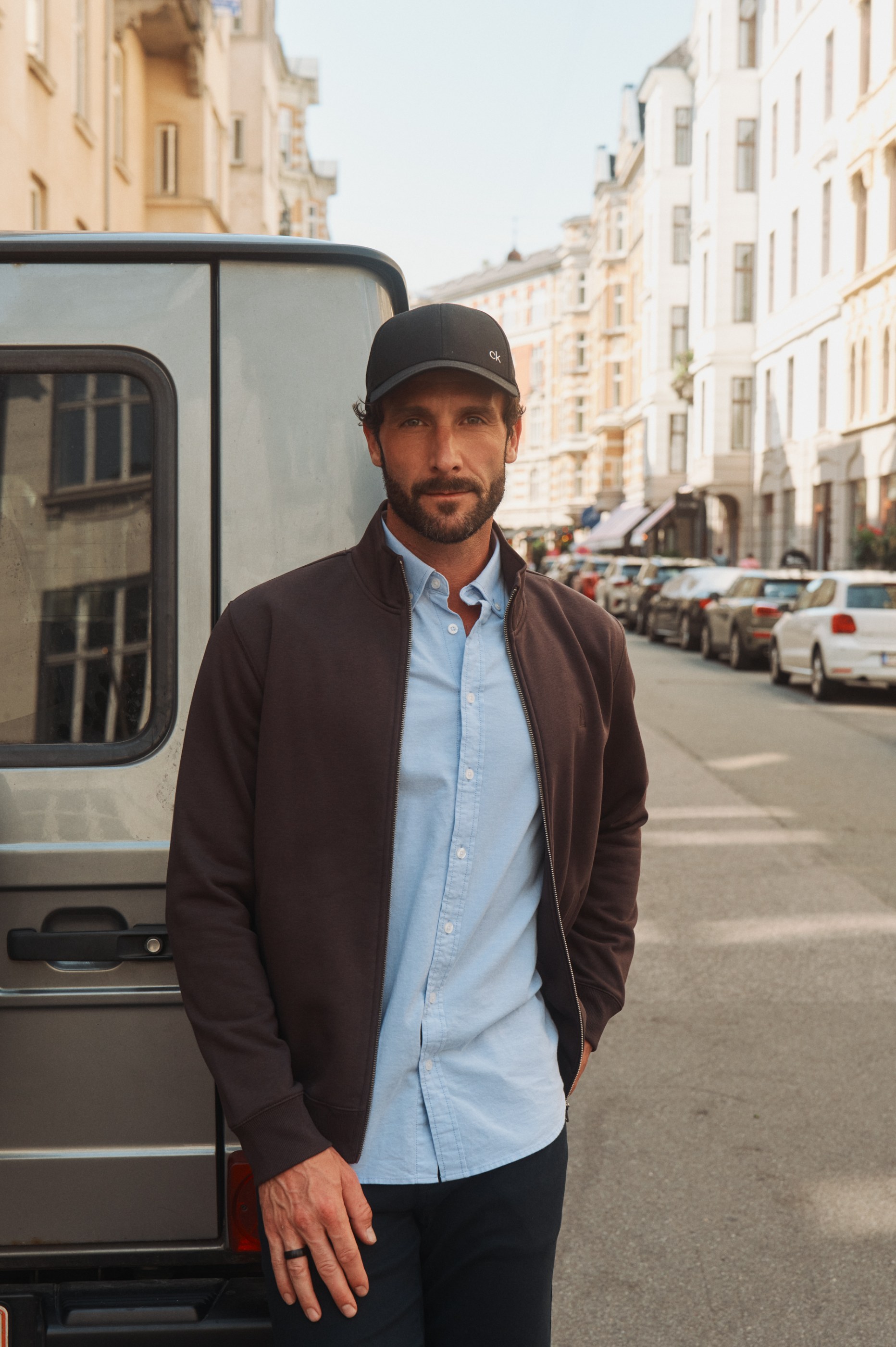 Bearded man in "ck" cap, light blue shirt, and brown jacket, standing on a sunny city street.