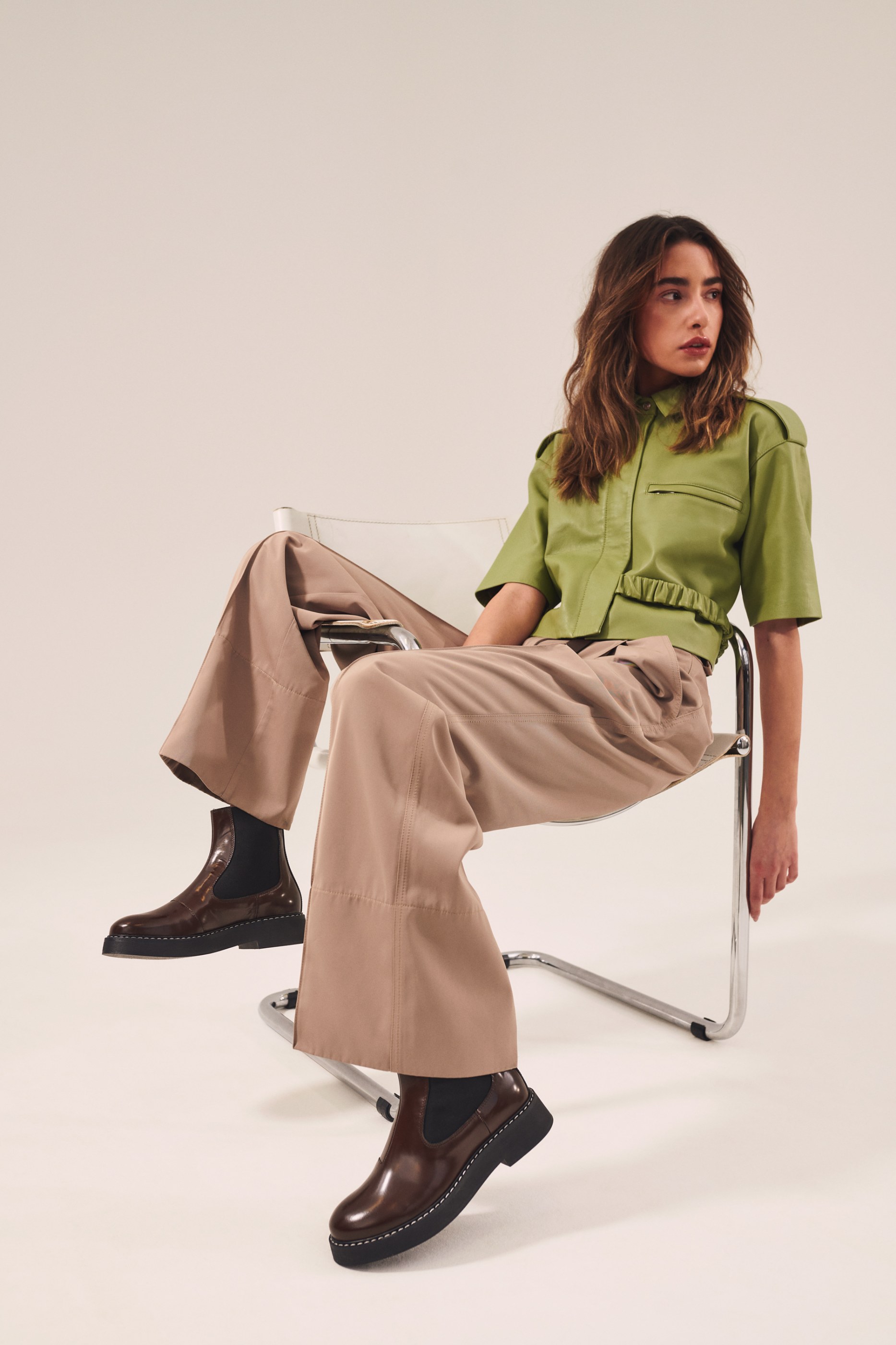 Woman wearing a green short-sleeved top, wide beige pants, and brown boots, seated on a chair.