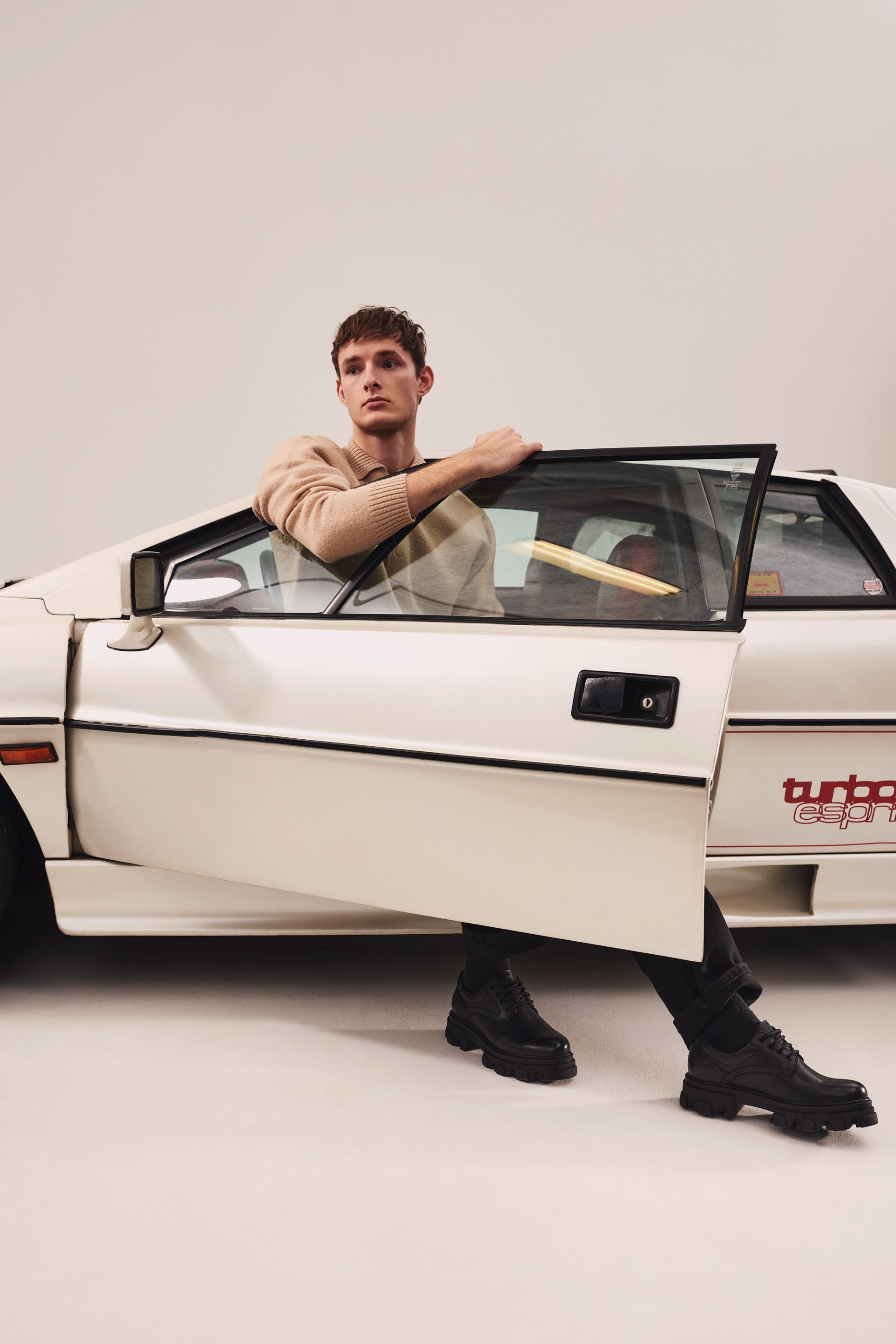 Young man in a beige sweater and black shoes leaning out of a white 'turbo esprit' car.