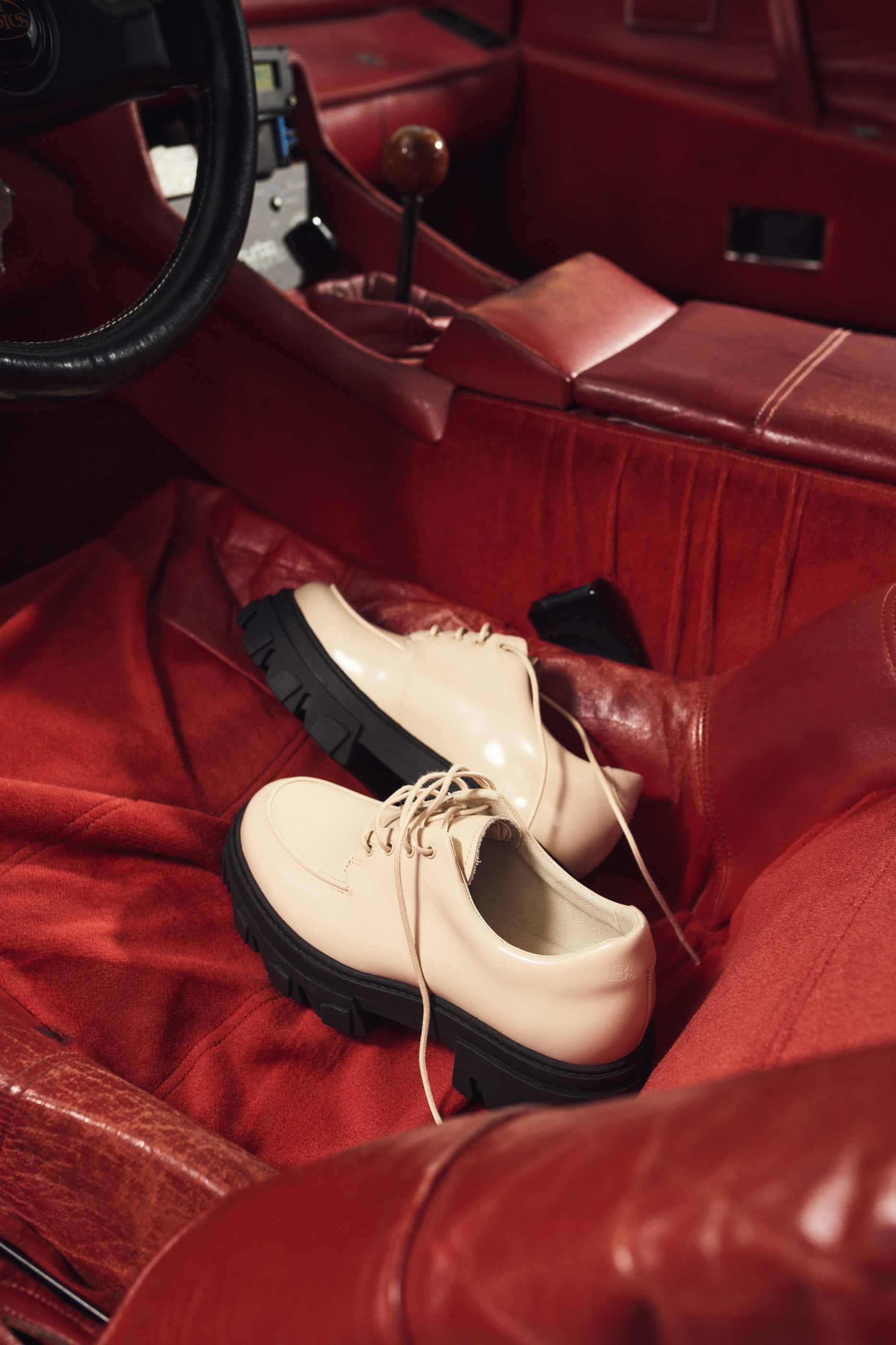 Two beige lace-up shoes with chunky black soles in a red car interior.