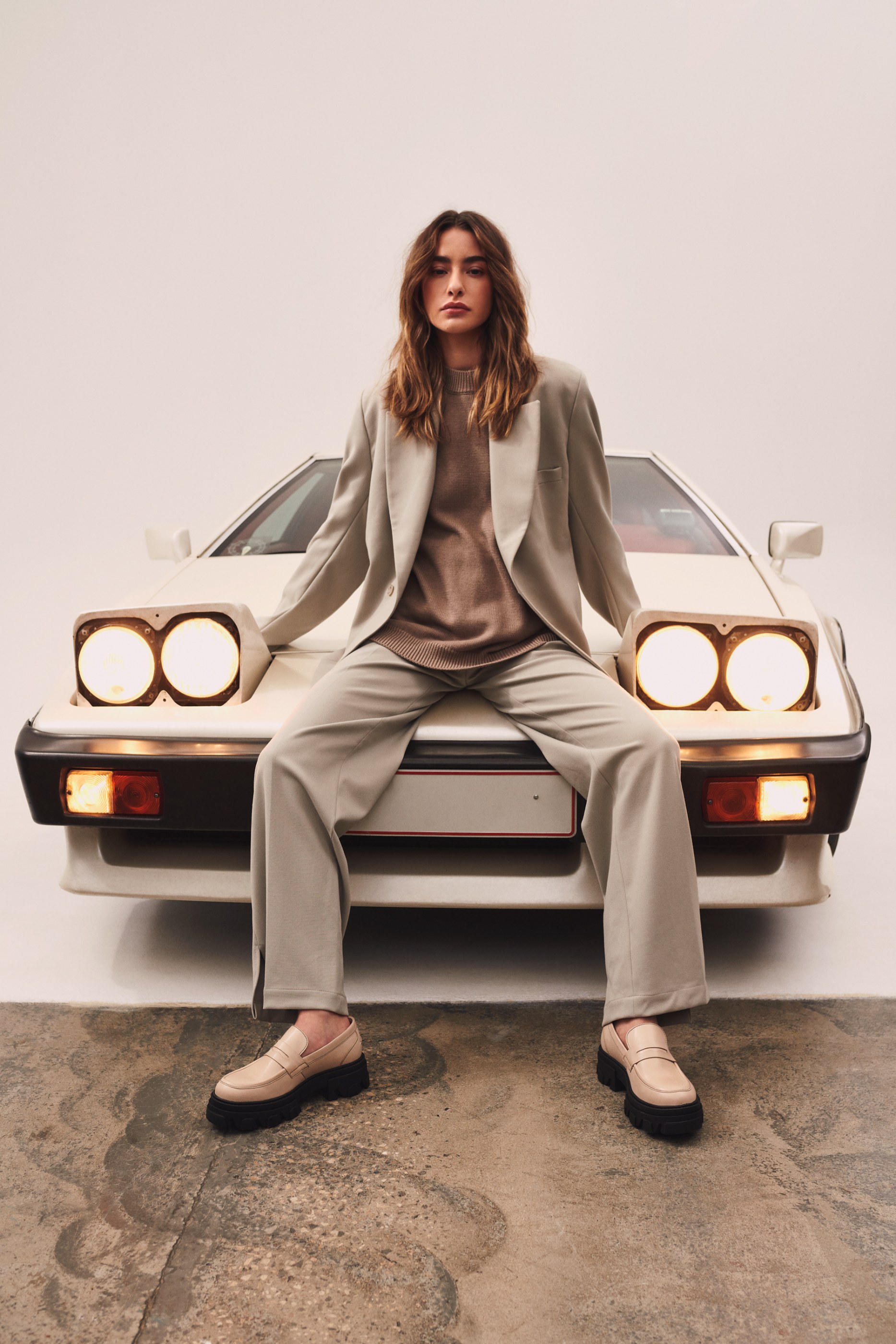 Woman in a neutral-toned suit and loafers on a white car with headlights on.