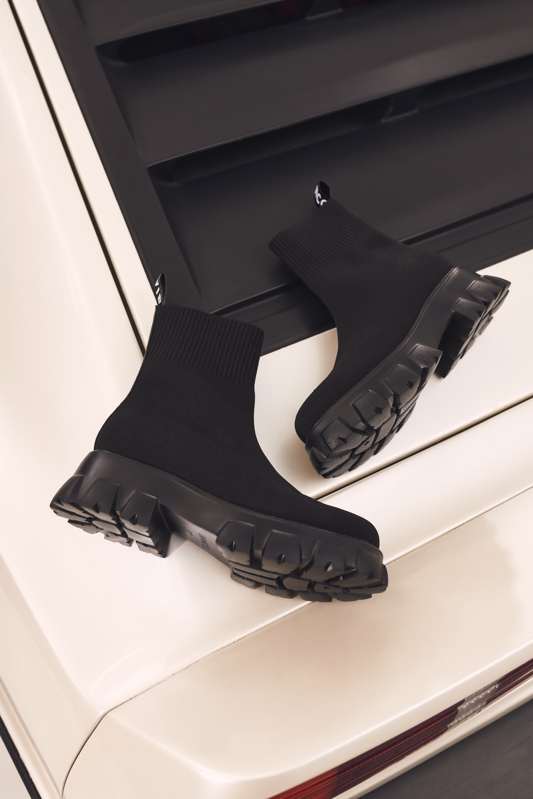 Two black knit sock boots with lug soles on a glossy white and black surface.