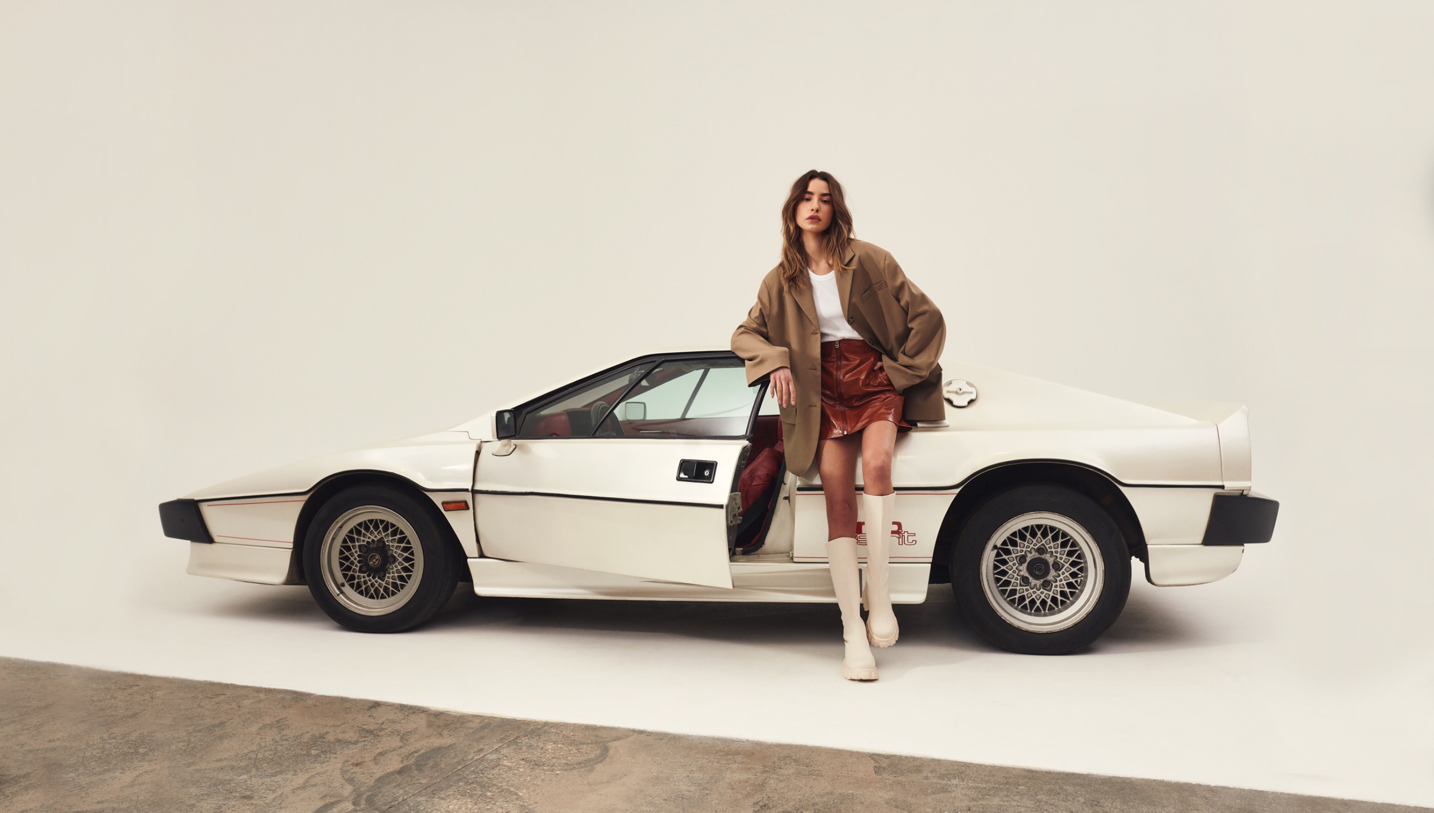 A stylish woman in a brown blazer and red leather skirt poses with a white vintage sports car.