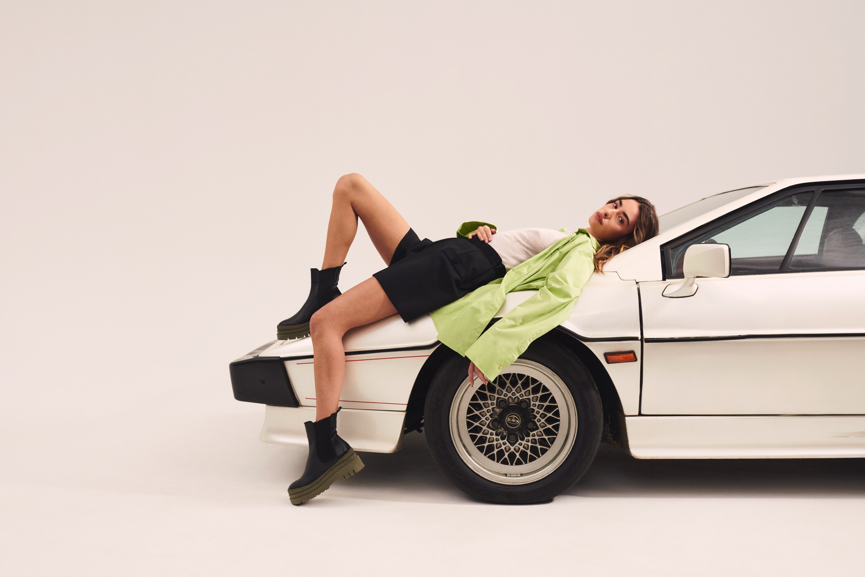 Woman in green shirt, black shorts, and boots reclines on a white sports car.